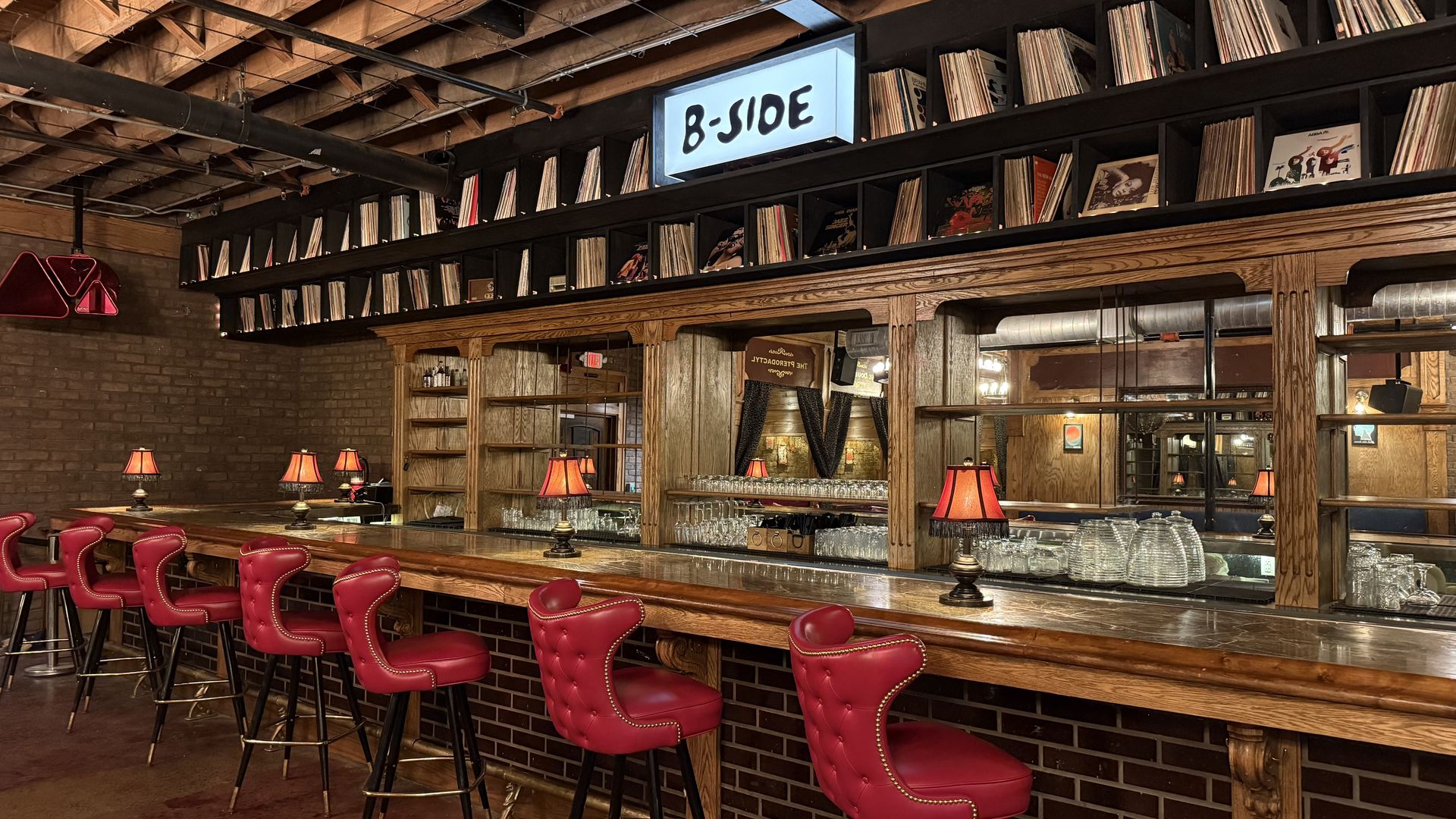 Bar interior with a long wooden counter, red tufted stools, brick walls, and vinyl shelves. A glowing sign reads "B-JOE" above the shelves; exposed ceiling beams and pipes.