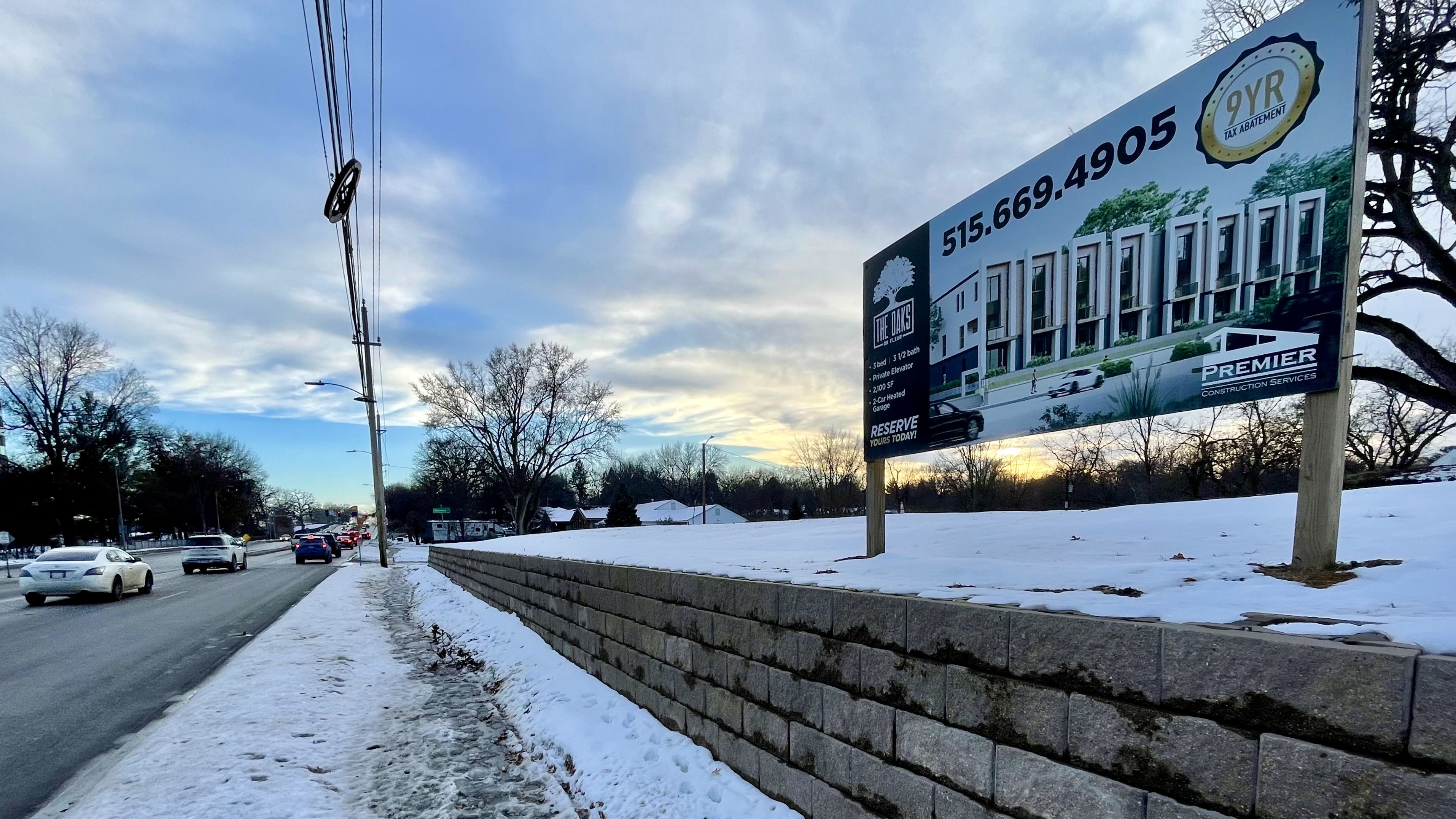 Snow-covered sidewalk and street with cars, a stone wall, leafless trees, and a large billboard advertising townhomes called The Oaks with phone number 515.669.4905 at sunset.