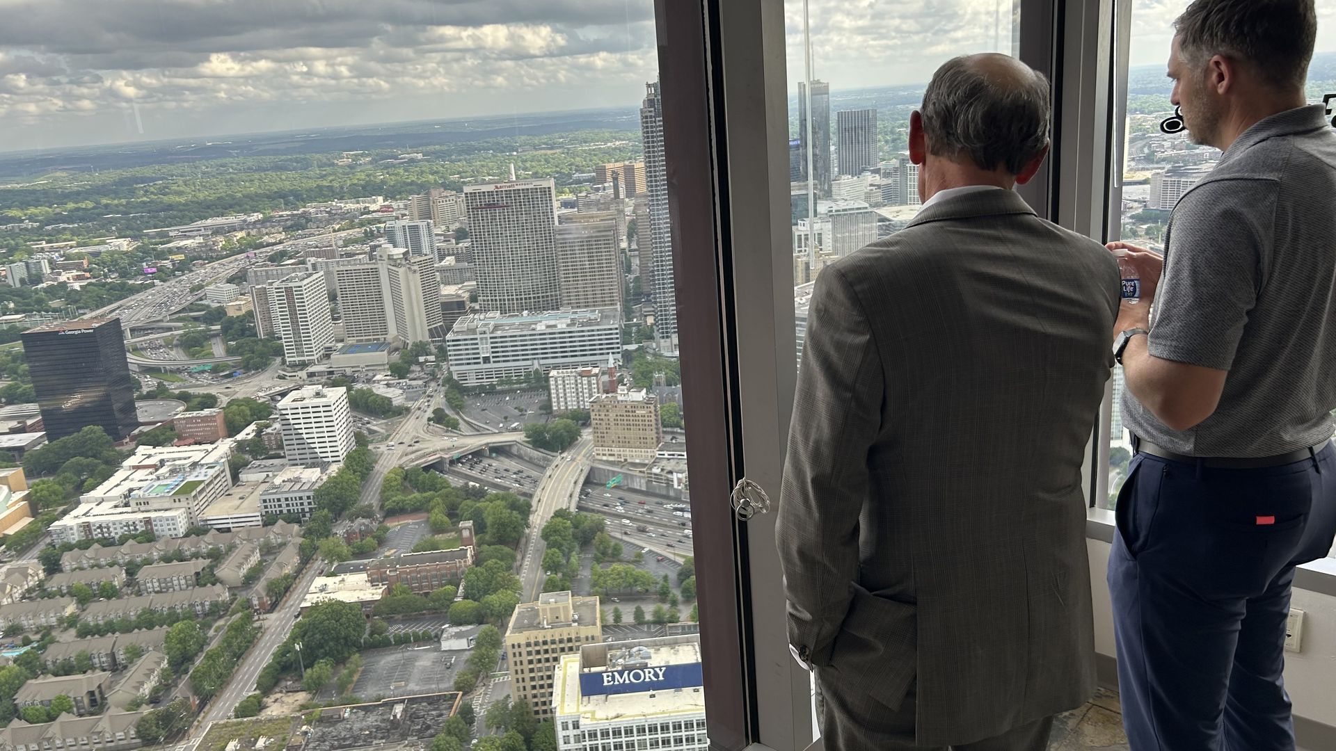 Alt-text: Two men look out a window in a skyscraper at an interstate weaving through a dense downtown area.