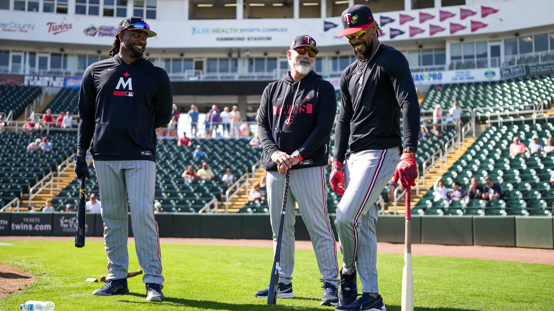 Three male baseball players stand on a sunlit field near the stands, smiling. They wear black tops with gray pinstriped pants; two hold bats while the third rests his. Stadium stands in background.