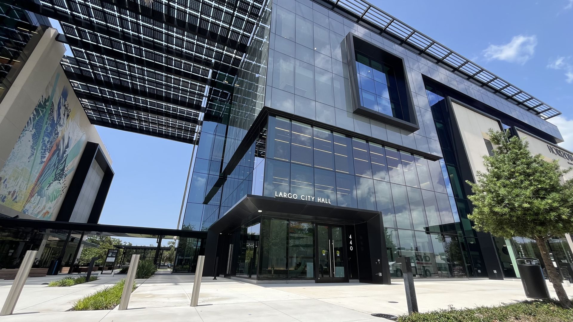 Low-angle view of a modern glass building with black metal frames and protruding square windows. The sign reads "Largo City Hall." A mural is on the left, a tree on the right, and a blue sky above.