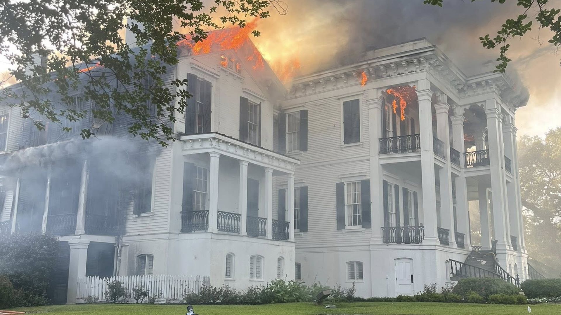 A large white mansion is shown smoking and on fire.