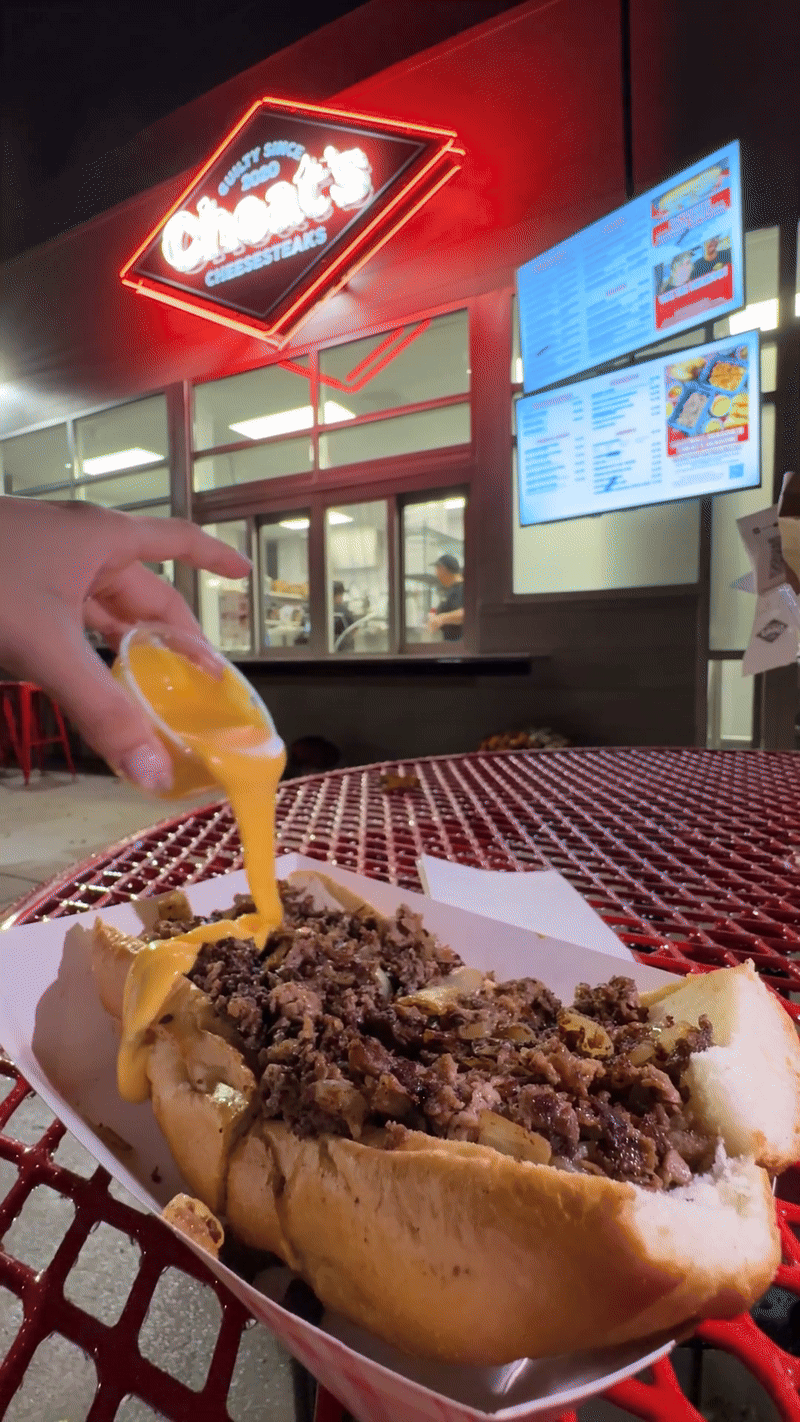 hand pouring cup of melted cheese onto cheesesteak