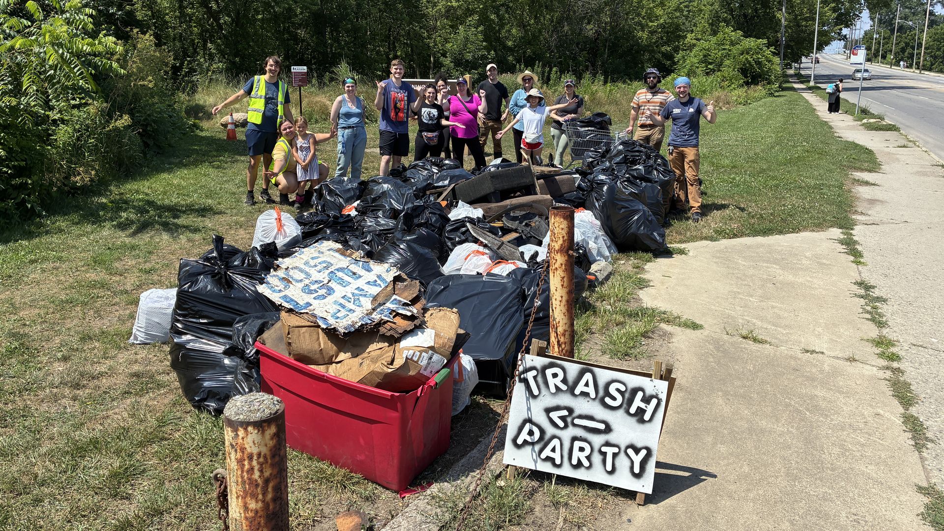 Group of volunteers posing behind large piles of black trash bags and debris outdoors near trees and a road, with a sign reading "Trash Party" on a sunny day.