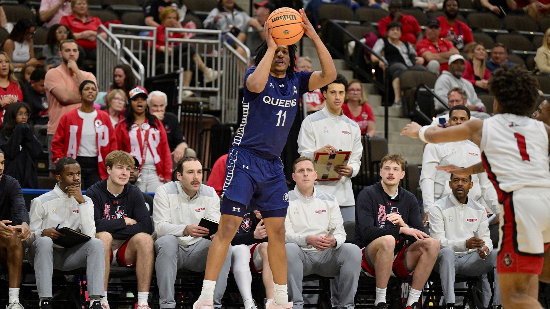 A basketball player in a blue Queens jersey (#11) leaps to shoot, while a defender in white and red guards him. Bench players and spectators watch from the sidelines.