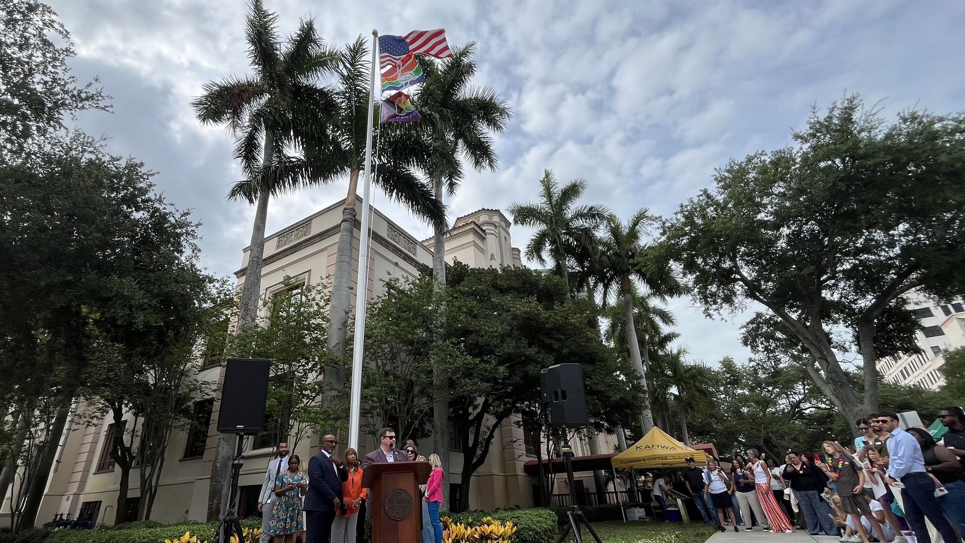 Public ceremony outside a beige building with palm trees. A flagpole flies the U.S. flag, a rainbow Pride flag, and a city of St. Pete flag. People stand near a podium; a yellow tent backs the crowd.