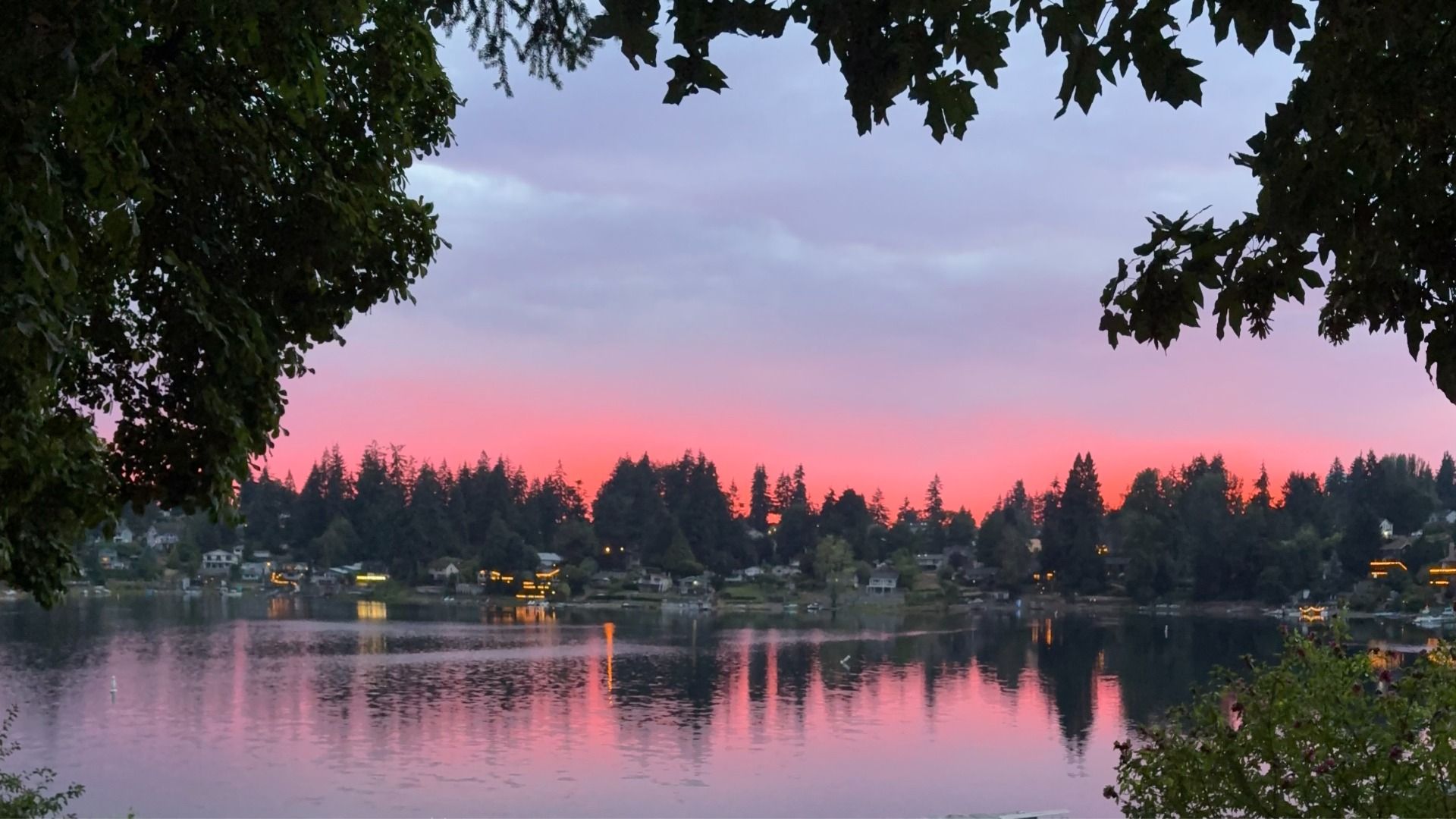 Pink and orange sunset light glows beneath a layer of smoky clouds over a calm lake, with tree silhouettes, waterfront homes and reflections shimmering on the water.