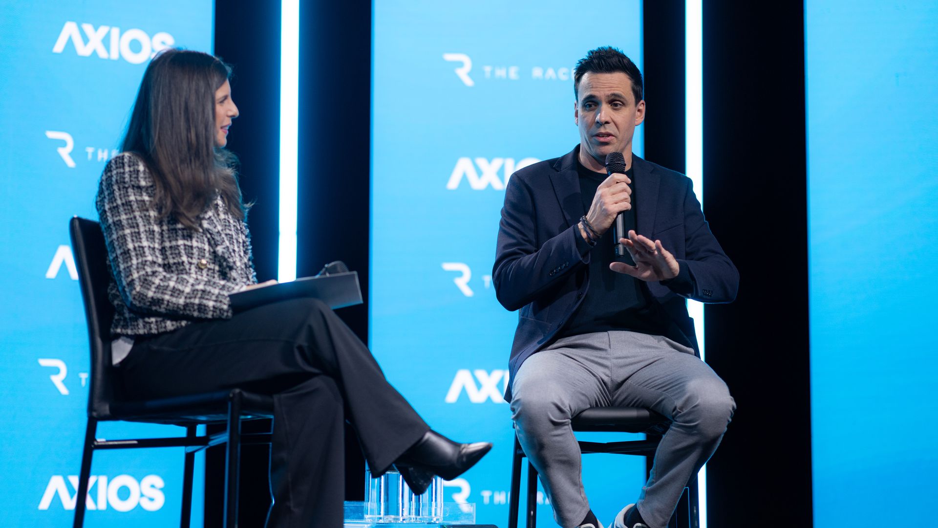 Two people seated on stage during an interview or discussion event. The man on the right speaks into a microphone; the woman on the left listens attentively. Blue screens behind show "AXIOS" and "THE RACE" logos.