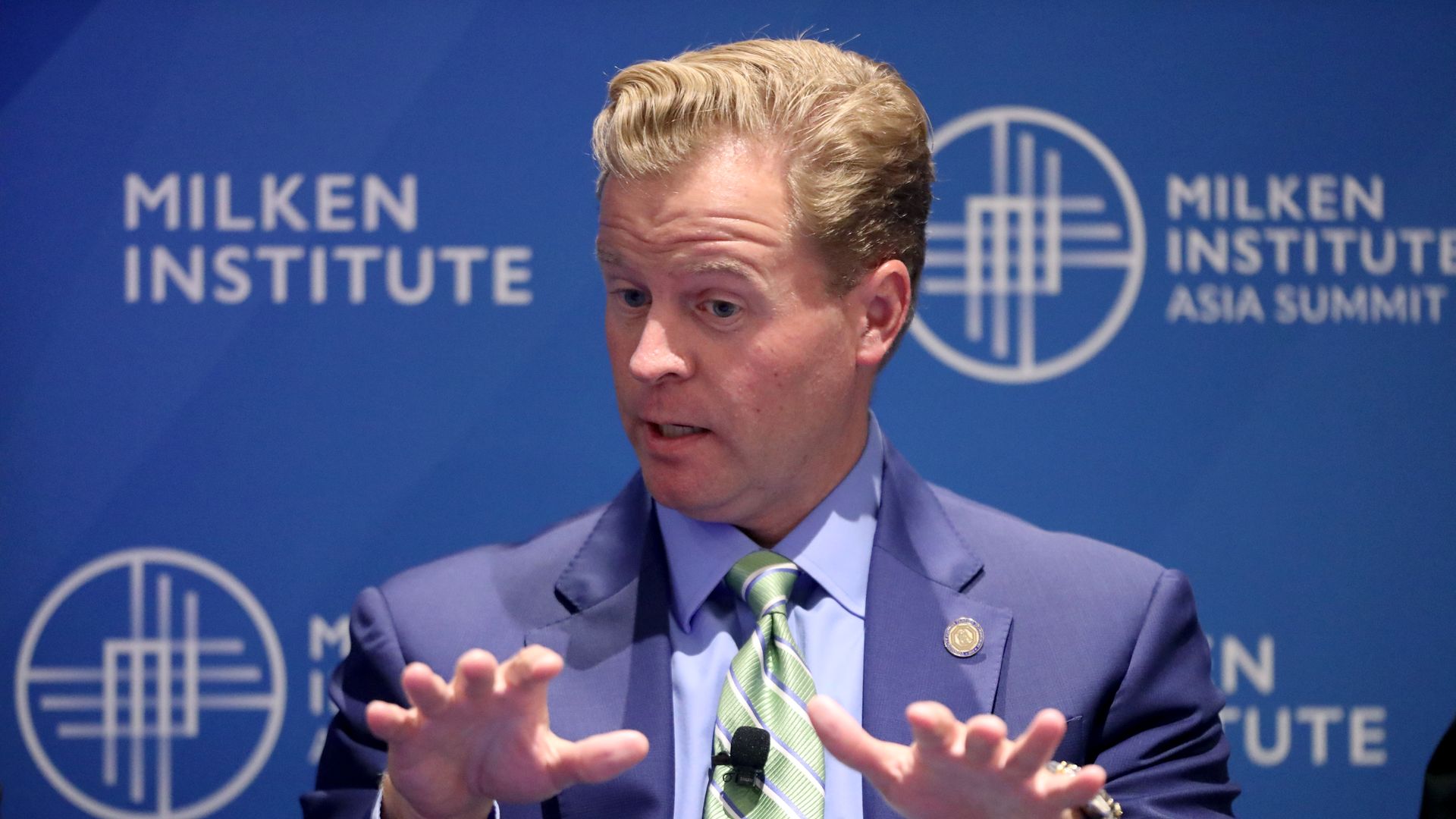 A man gesturing with both hands, seen from the shoulders up in a blue suit in an official looking setting. Milken Institute written on the walls behind him. 