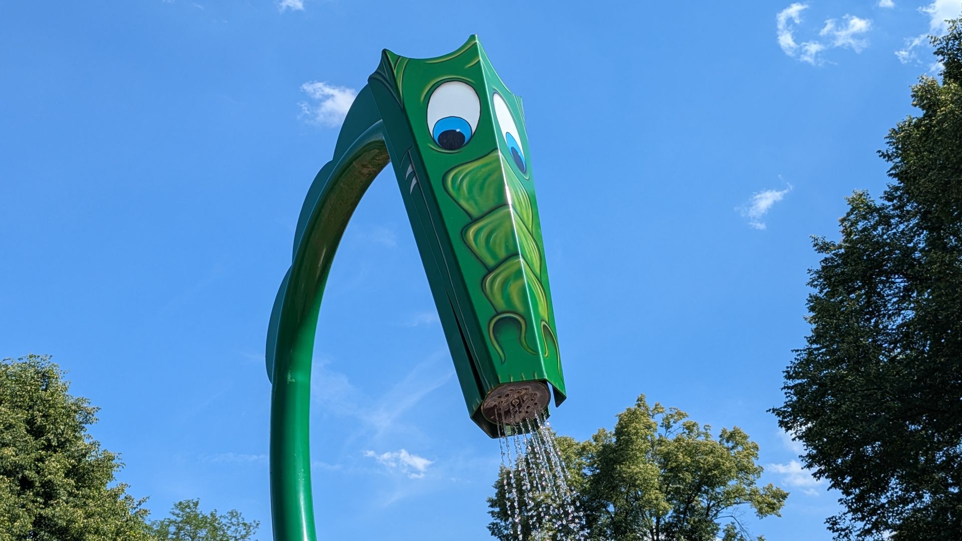 Green, curved water slide shaped like a cartoon monster with large eyes, towering against a bright blue sky, with water dripping from its base and trees in the background.