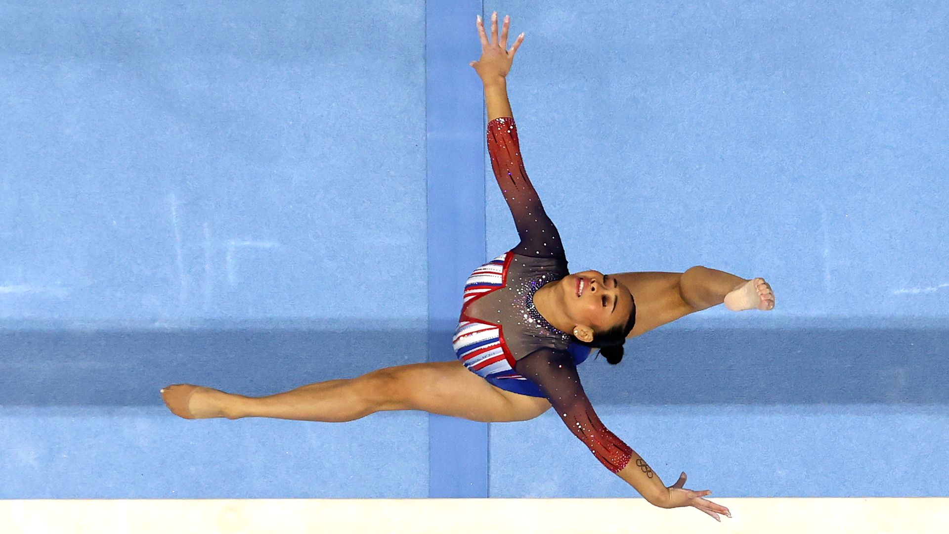 A gymnast in red white and blue leotard photographed from above mid-leap, with their head thrown back, their arms extended and their legs raised