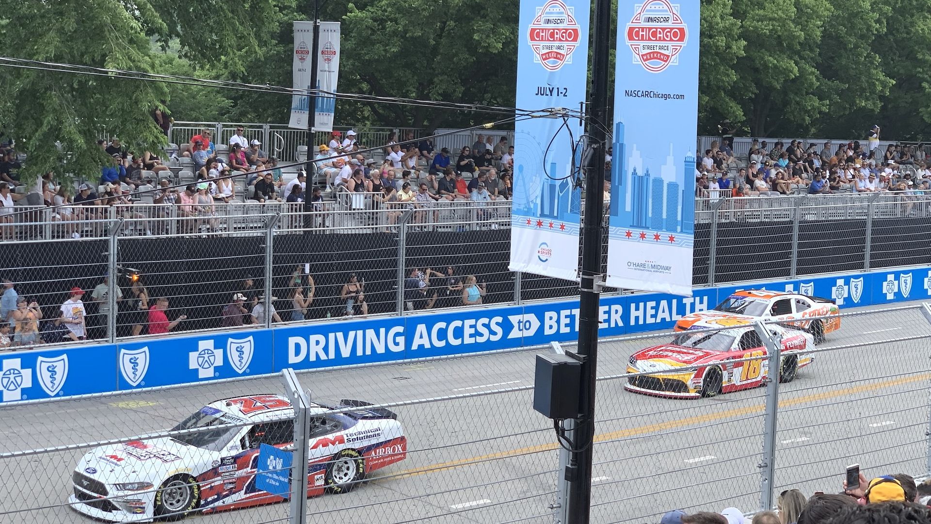 Crowd sits in grandstands beside a racetrack, under tall blue Chicago banners for a NASCAR weekend. Green trees in background; race cars on the track behind a chain-link fence.