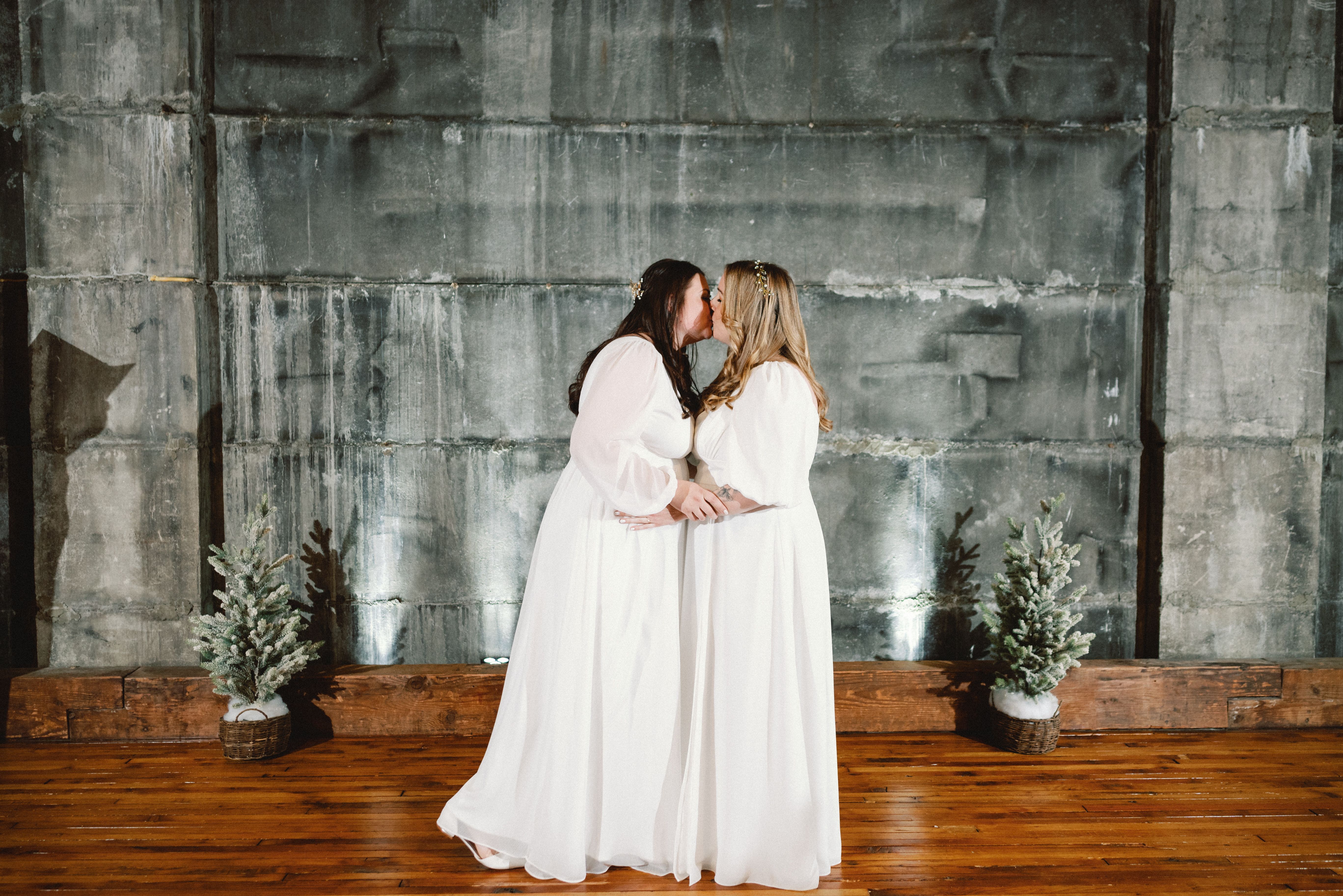 Two brides in white kiss as they wed at Olio, an industrial wedding venue in Peabody, Massachusetts.