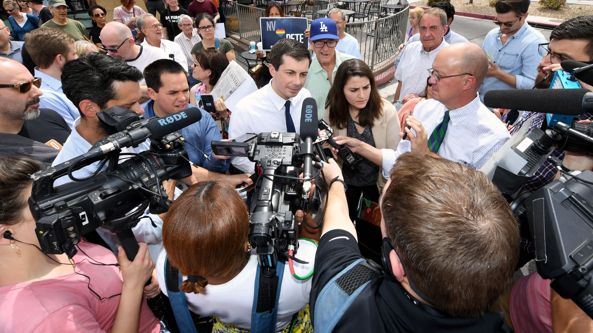 In this image, Pete Buttigieg stands in the center of a crowded ring of reporters who hold microphones towards him.