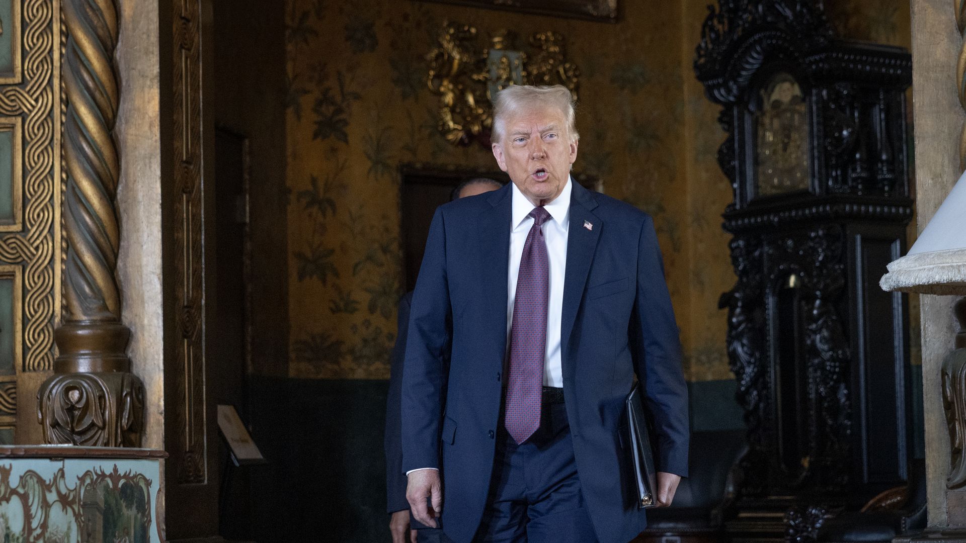 President Trump in a navy suit, white shirt, and purple tie walks through a gilded, ornate interior, carrying a black folder; an American flag lapel pin is visible.