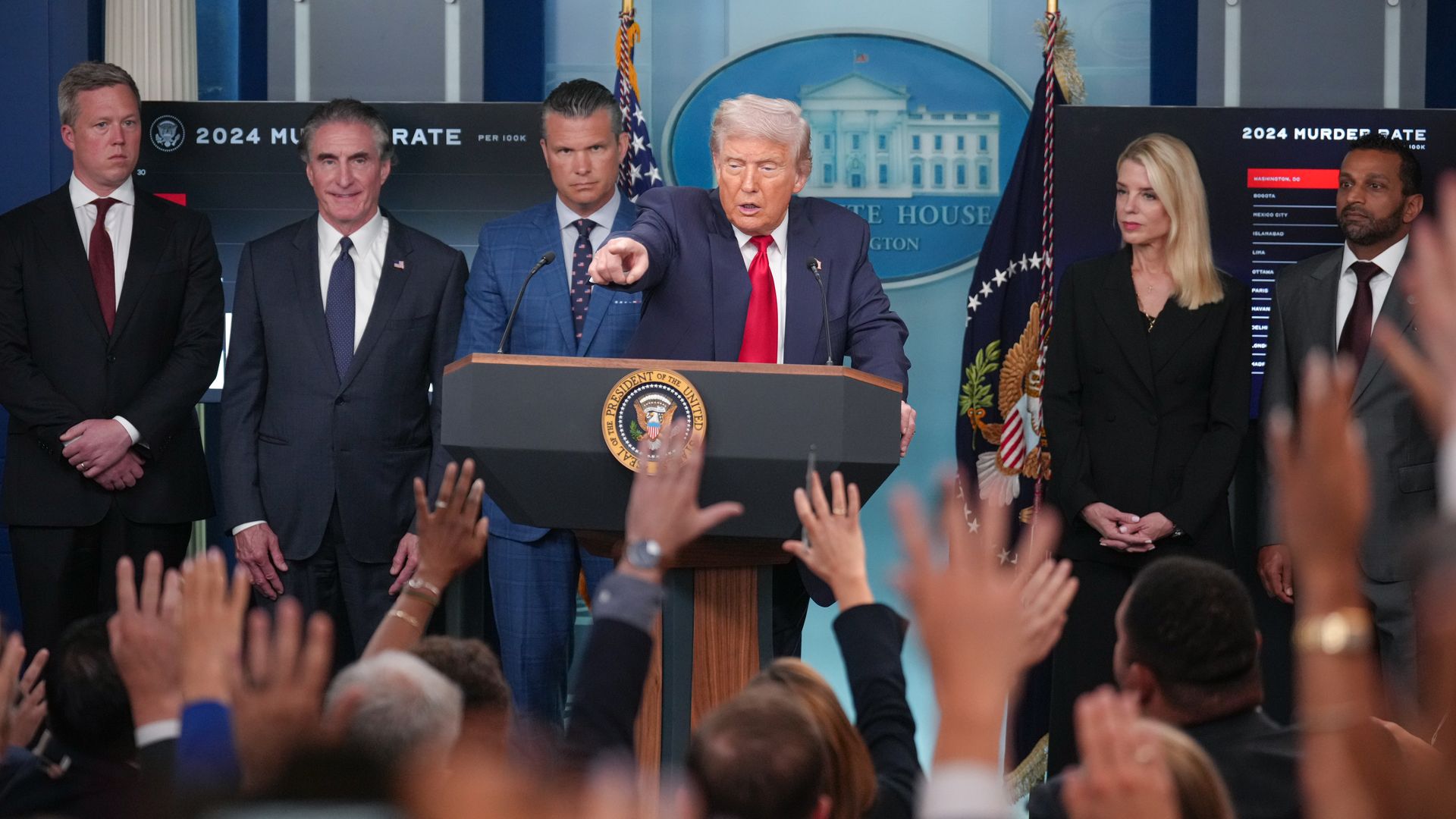 Donald Trump speaking at White House podium with five people behind him, audience raising hands, 2024 murder rate charts visible on screens in background.