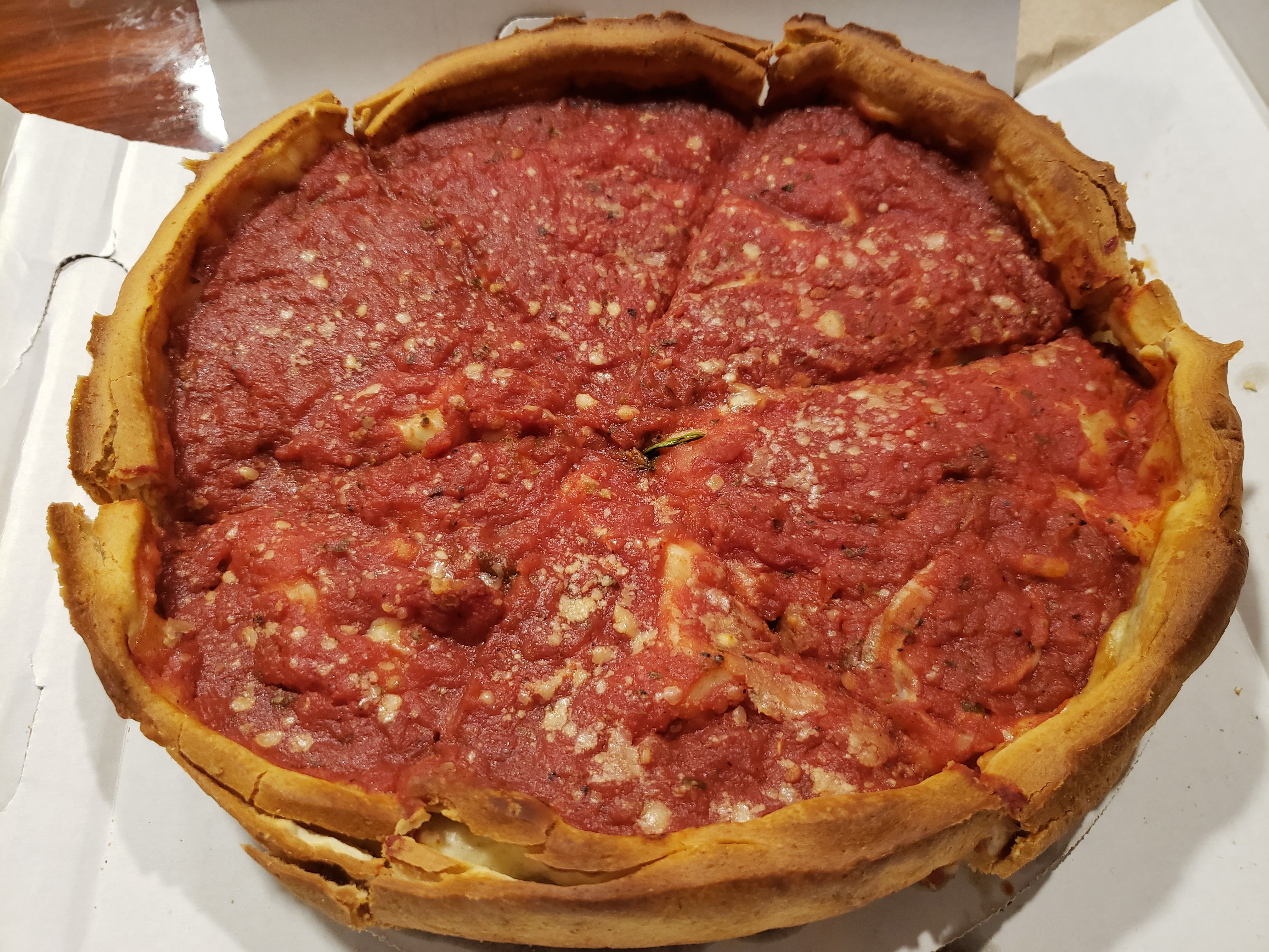 Deep-dish pizza sliced into six pieces with thick tomato sauce topping and golden-brown crust, inside a white pizza box on a wooden table.