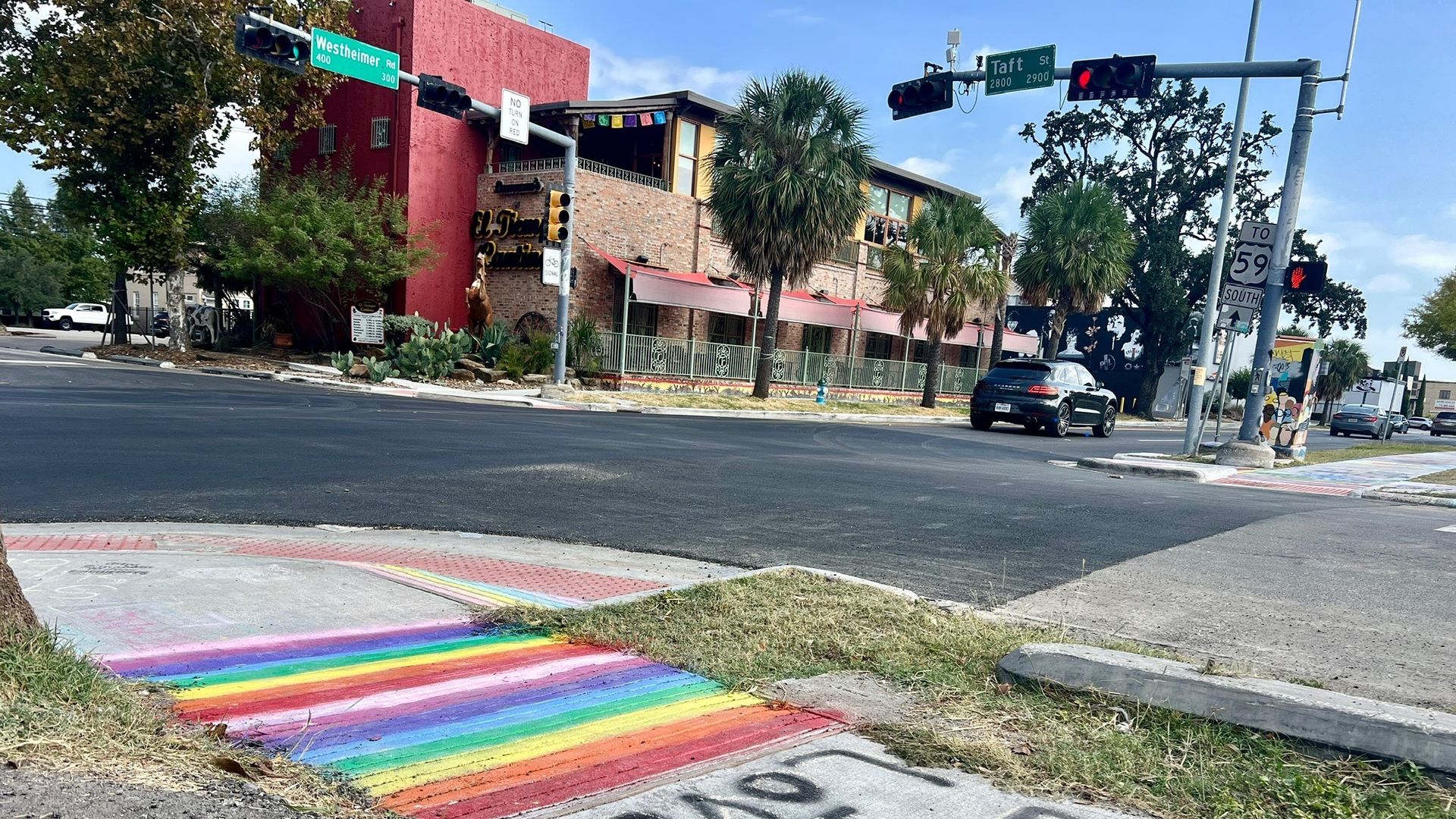 Street corner with a rainbow-painted crosswalk and graffiti on the sidewalk reading "LOVE & LOVE" next to a colorful fan-shaped design. Traffic lights and street signs for Westheimer Rd and Taft St visible.