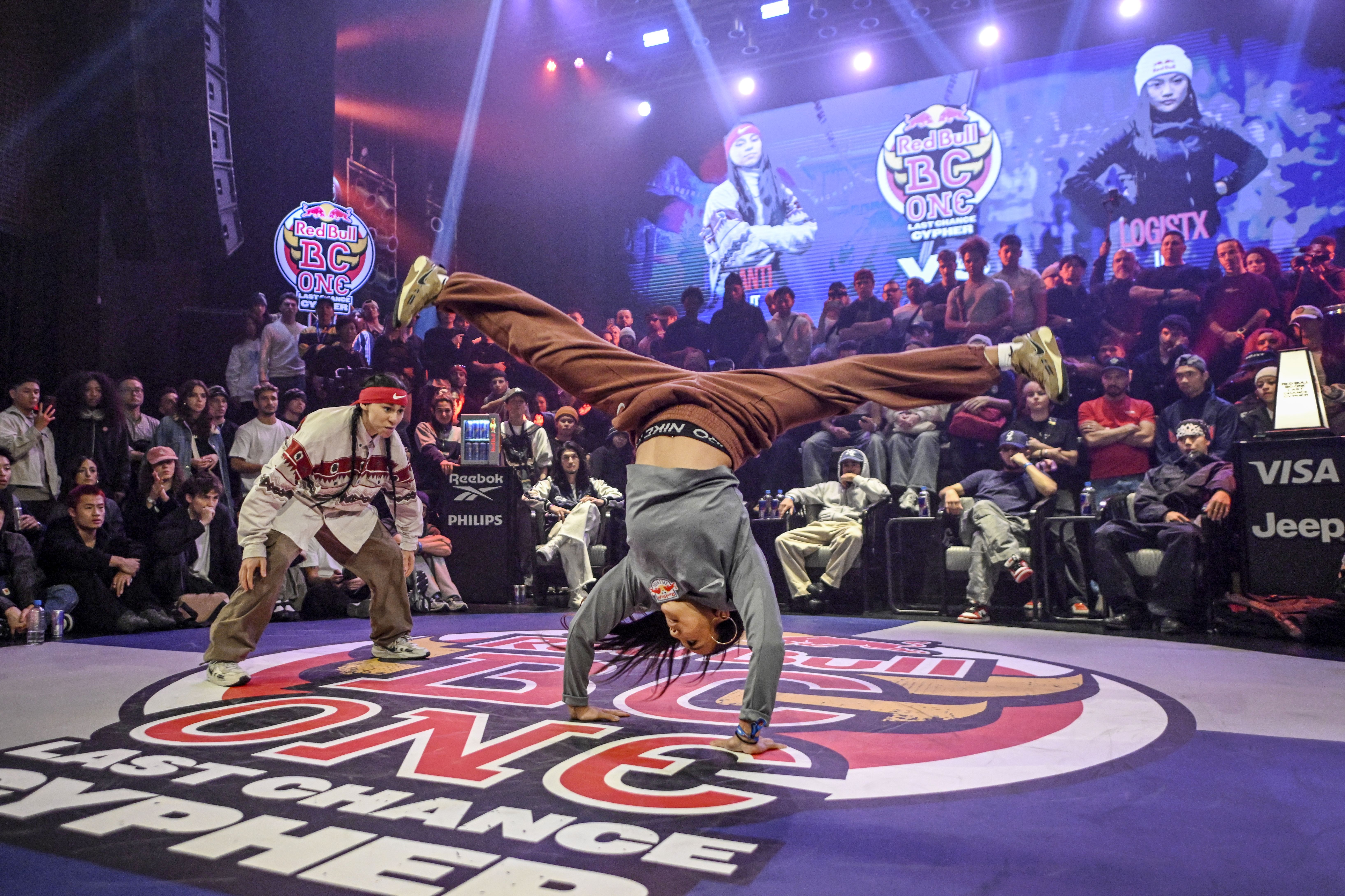 Two breakdancers perform on a circular stage under purple lighting as a crowded audience watches; large Red Bull BC One logos and a digital backdrop fill the scene.