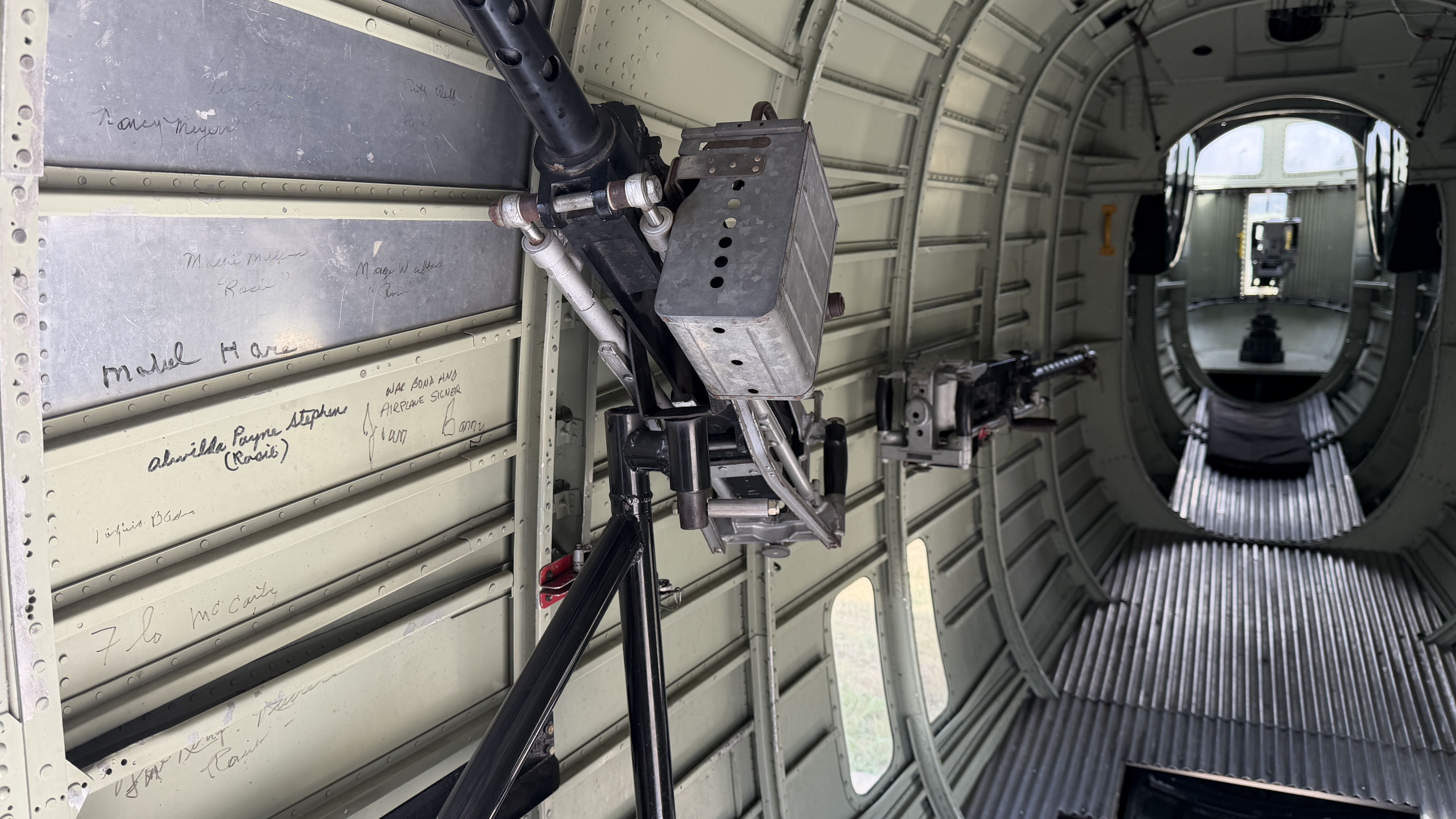 Interior of a vintage military aircraft with a mounted gun-like apparatus on a tripod, riveted gray walls with handwritten signatures, and a circular aft opening visible at the far end.