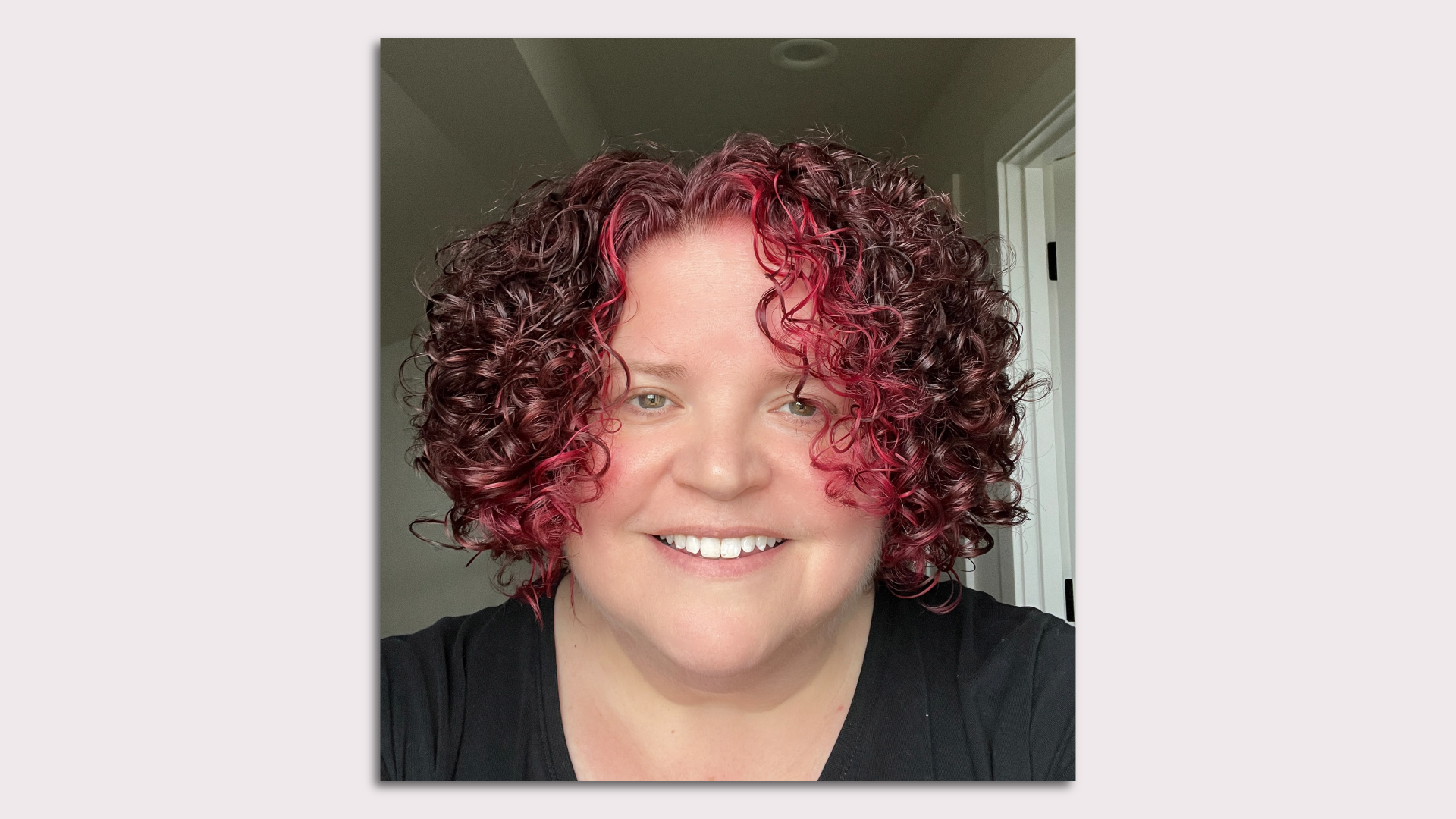 A smiling person with a mane of red and pink curls.
