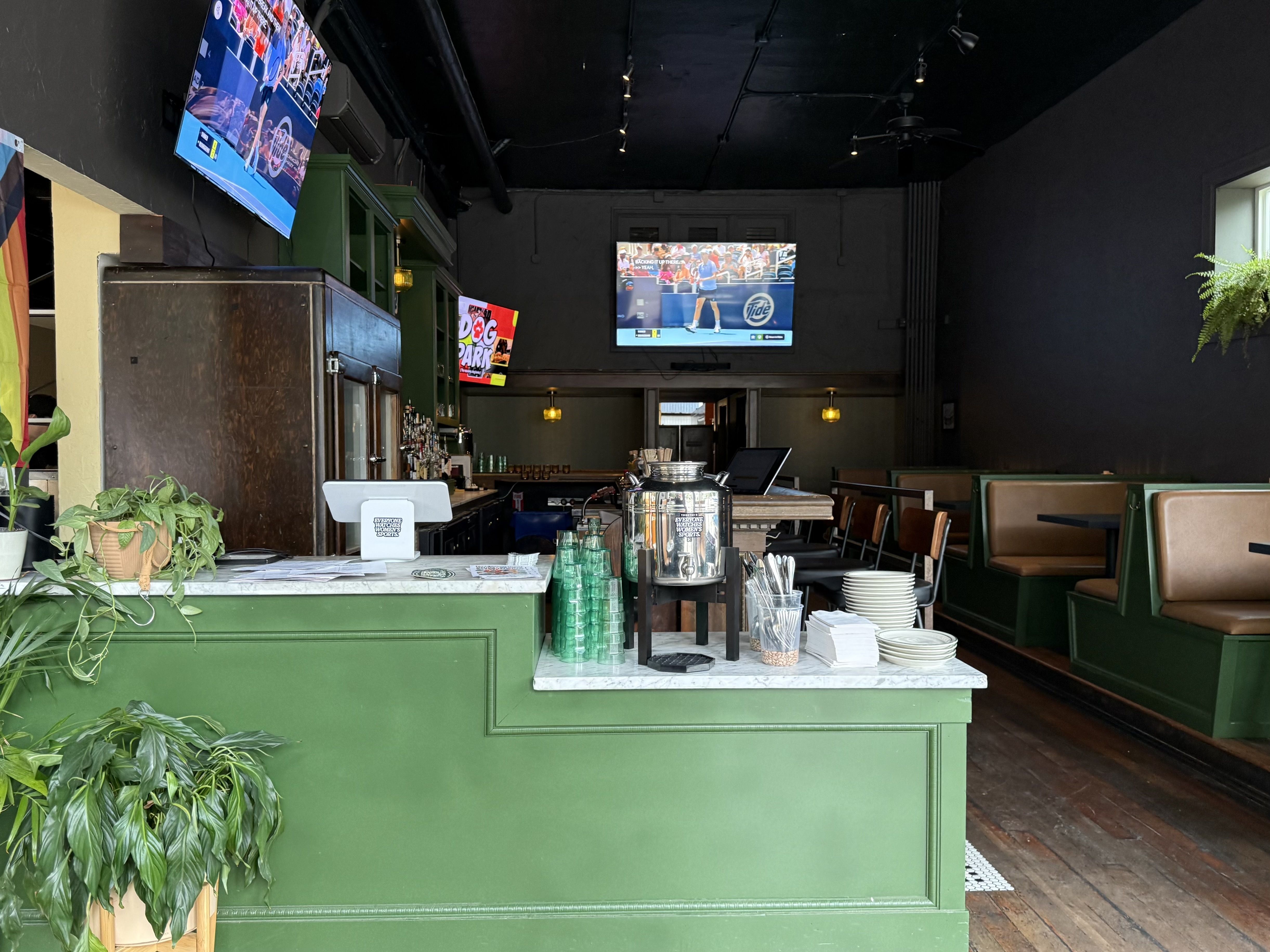 Interior of a bar with green counters and booths, a wooden floor, plants, and three TVs showing different programs including a tennis match and a sign for "Dog Park".
