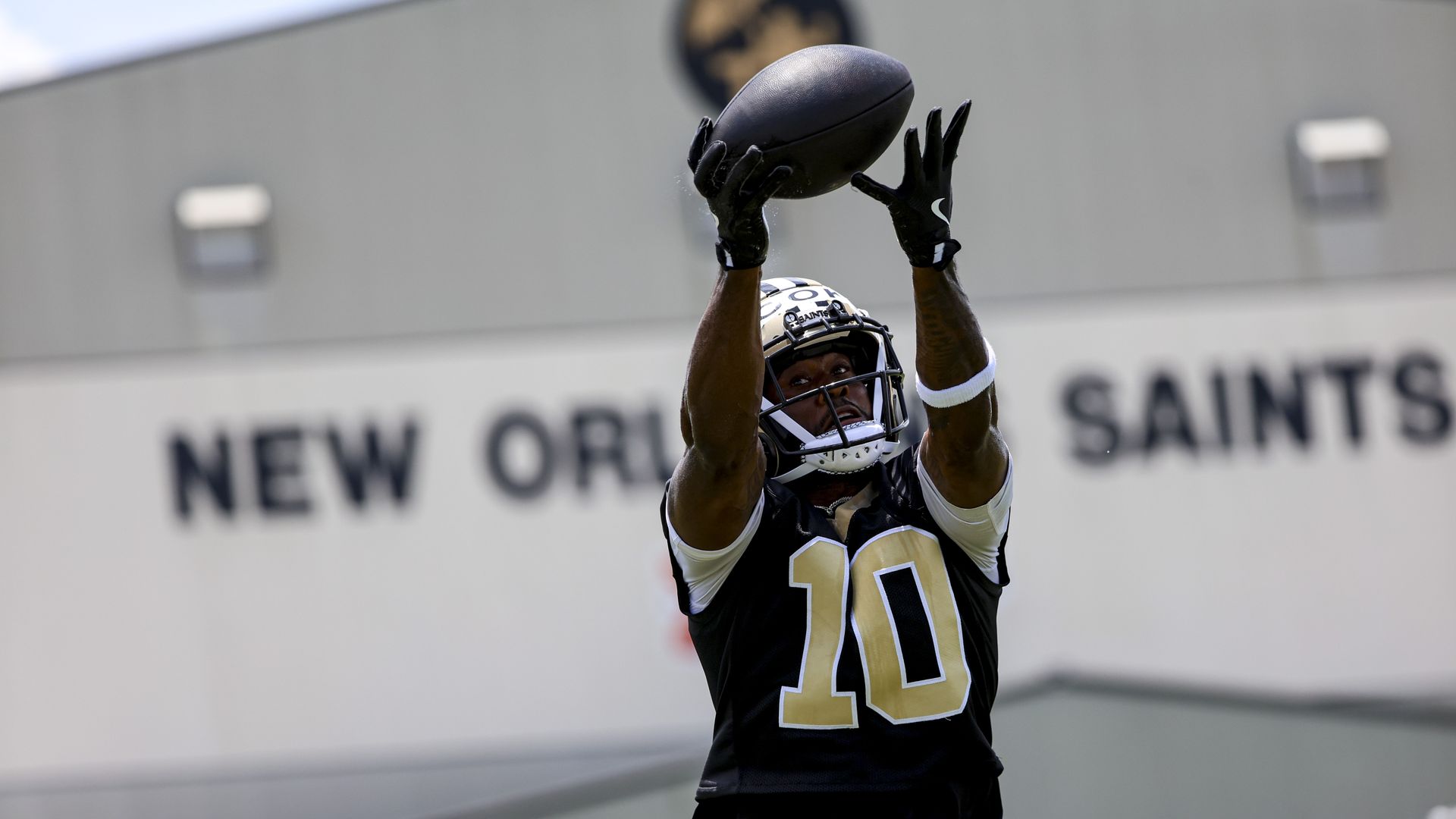 Football player wearing black New Orleans Saints jersey number 10 and helmet, catching a ball during practice outside a building with "New Orleans Saints" sign.