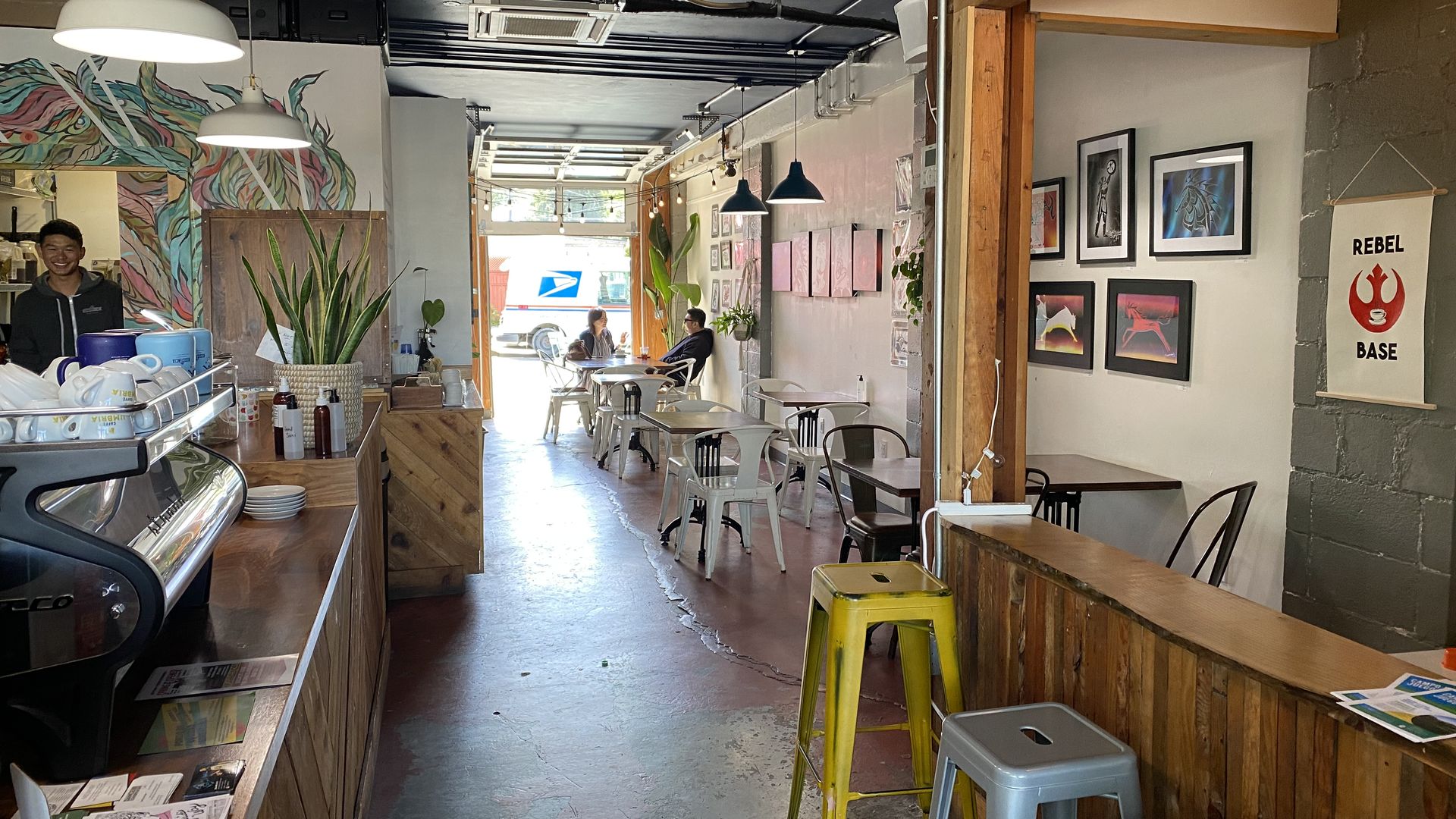 View of a coffee shop with art on the walls and a back wall that opens to the outside, with two people talking at a table. 
