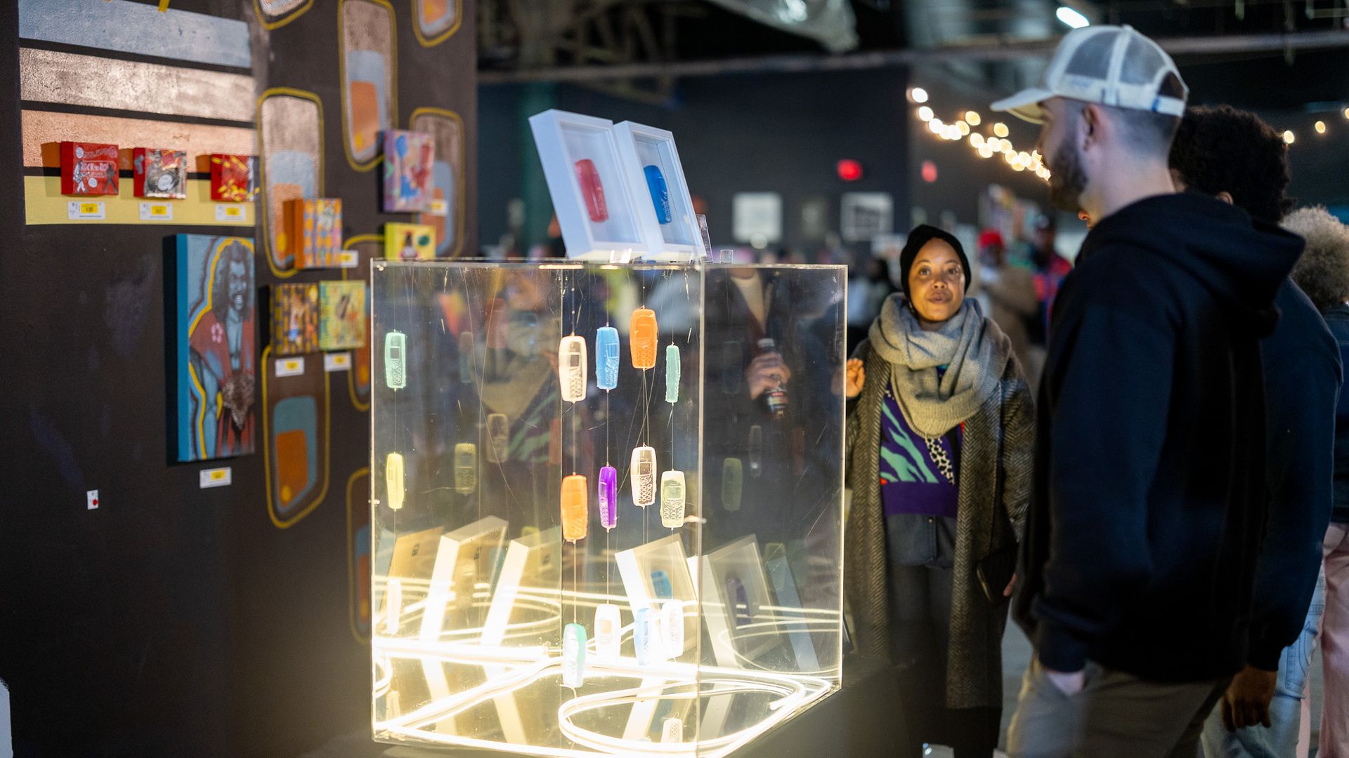 Gallery scene with illuminated display case featuring colorful hanging retro mobile phone models, visitors observe artwork on dark walls with bright string lights in the background.