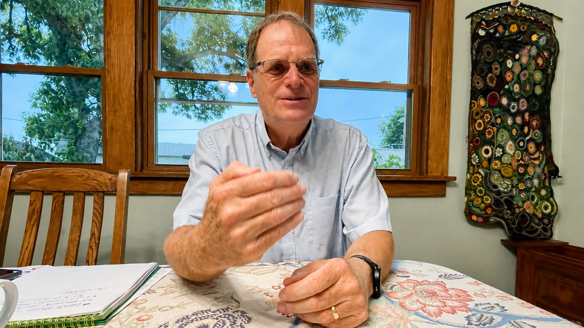 Man with glasses in a light-blue check shirt sits at a table with a floral tablecloth, hands clasped. Behind him are wooden windows with trees outside and a colorful quilt wall hanging.