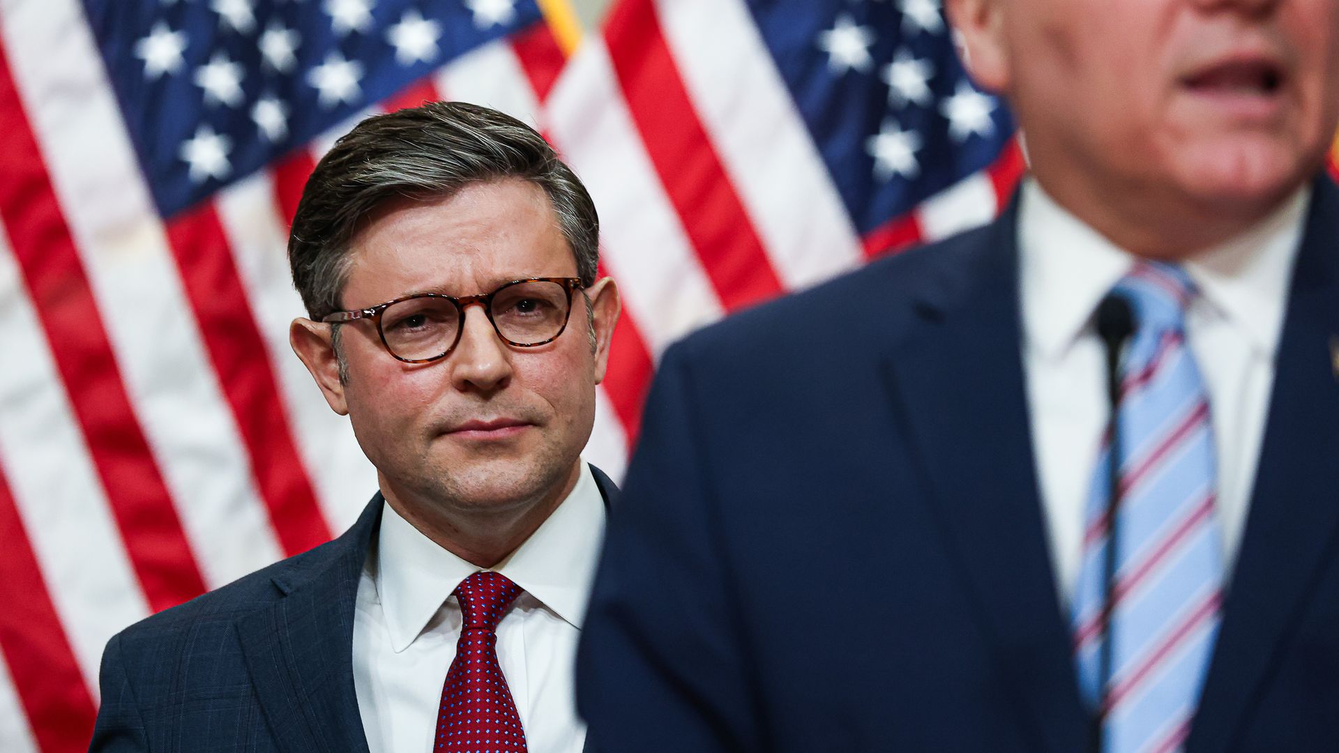House Speaker Mike Johnson, wearing a blue blazer, white shirt and red tie, stands behind a colleague and in front of a row of American flags.