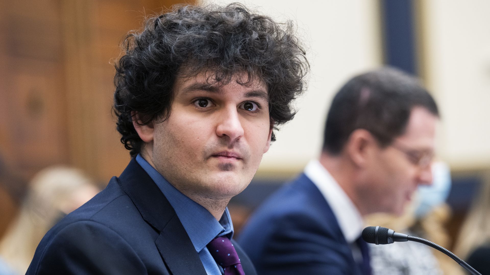 Sam Bankman-Fried looks toward a camera while seated inside a Congressional hearing room.