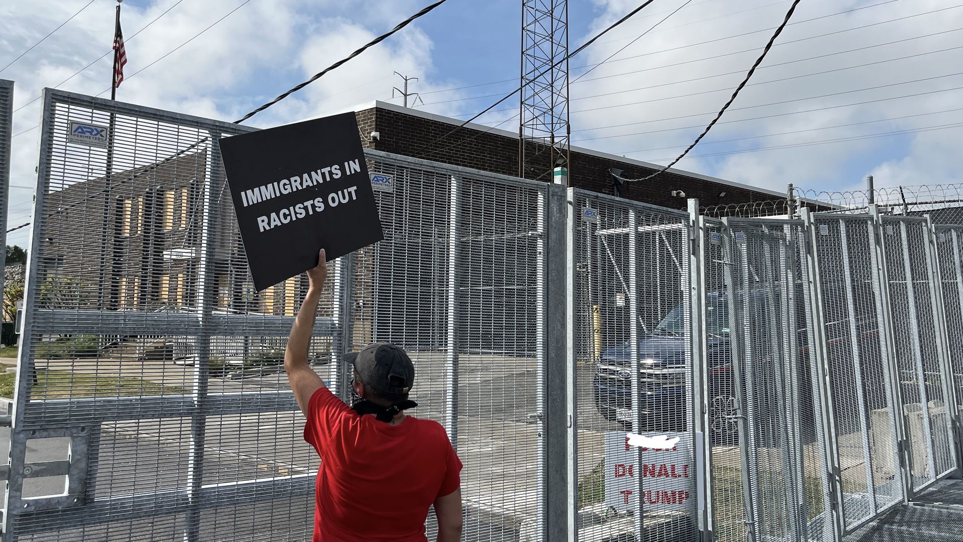 Person in a red shirt and cap holding a black sign reading "IMMIGRANTS IN RACISTS OUT" behind a tall metal fence topped with barbed wire on a partly cloudy day.