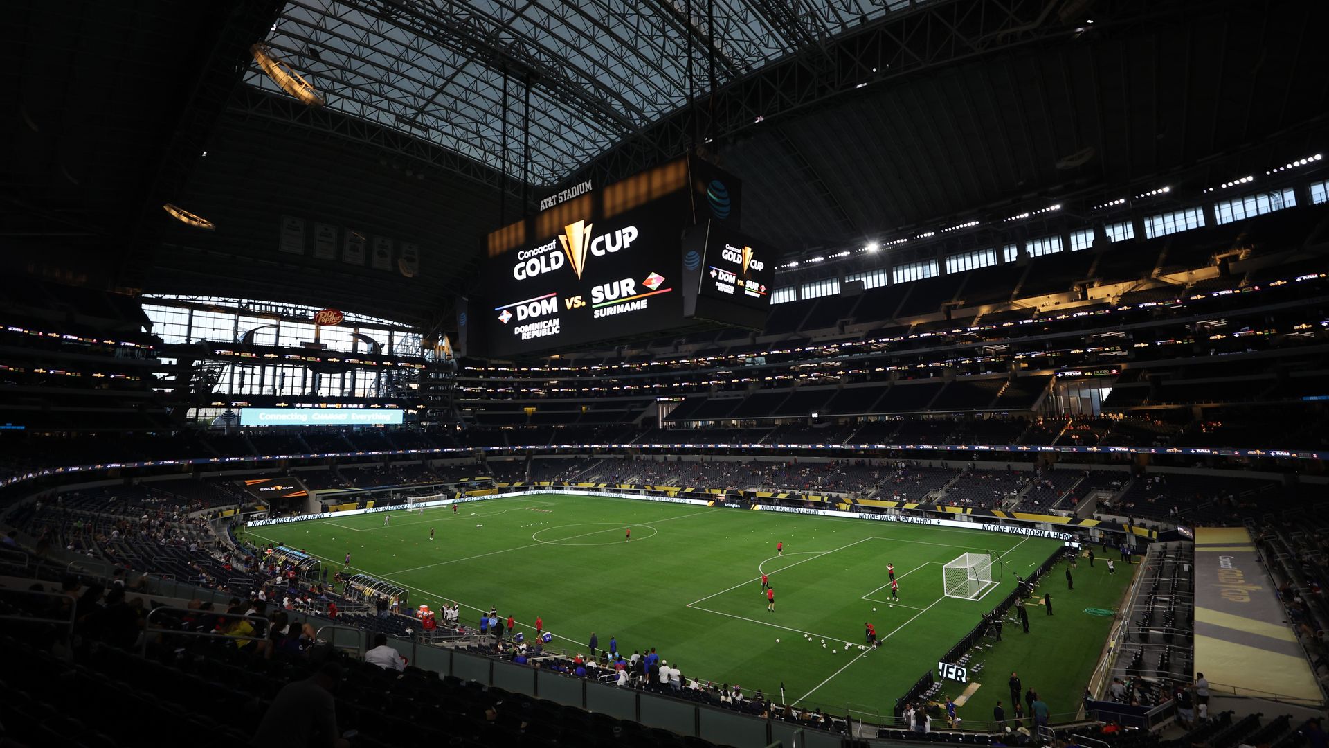 Interior view of AT&T Stadium with a soccer field and players warming up with the scoreboard displaying the Concacaf Gold Cup logo