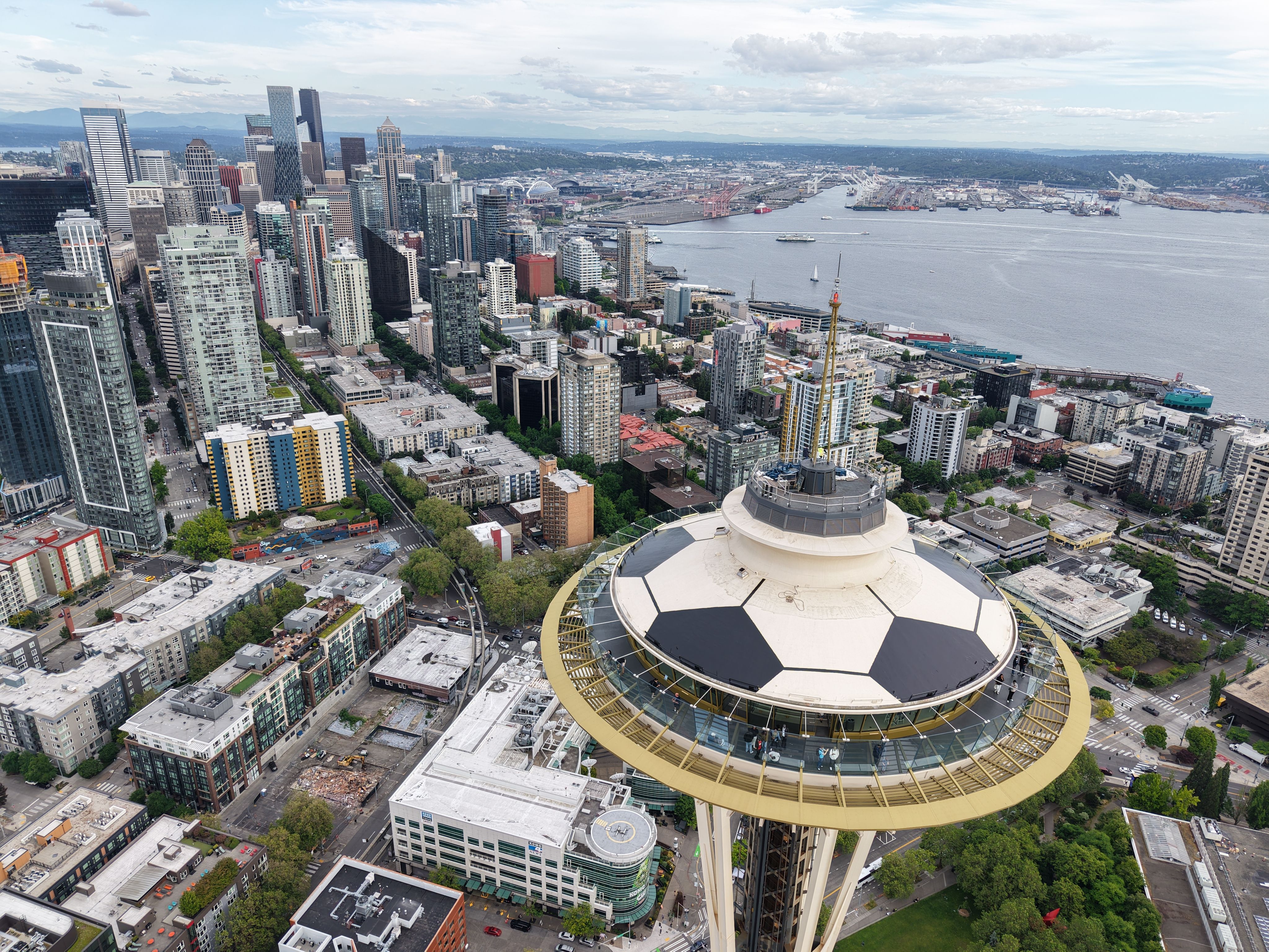 Panoramic view of downtown Seattle: tall skyscrapers on the left, Space Needle in the foreground, painted with black-and-white soccer ball pattern. In the distance are the waterfront with ships and cranes, low-rise blocks below, bay and distant hills under a cloudy sky.