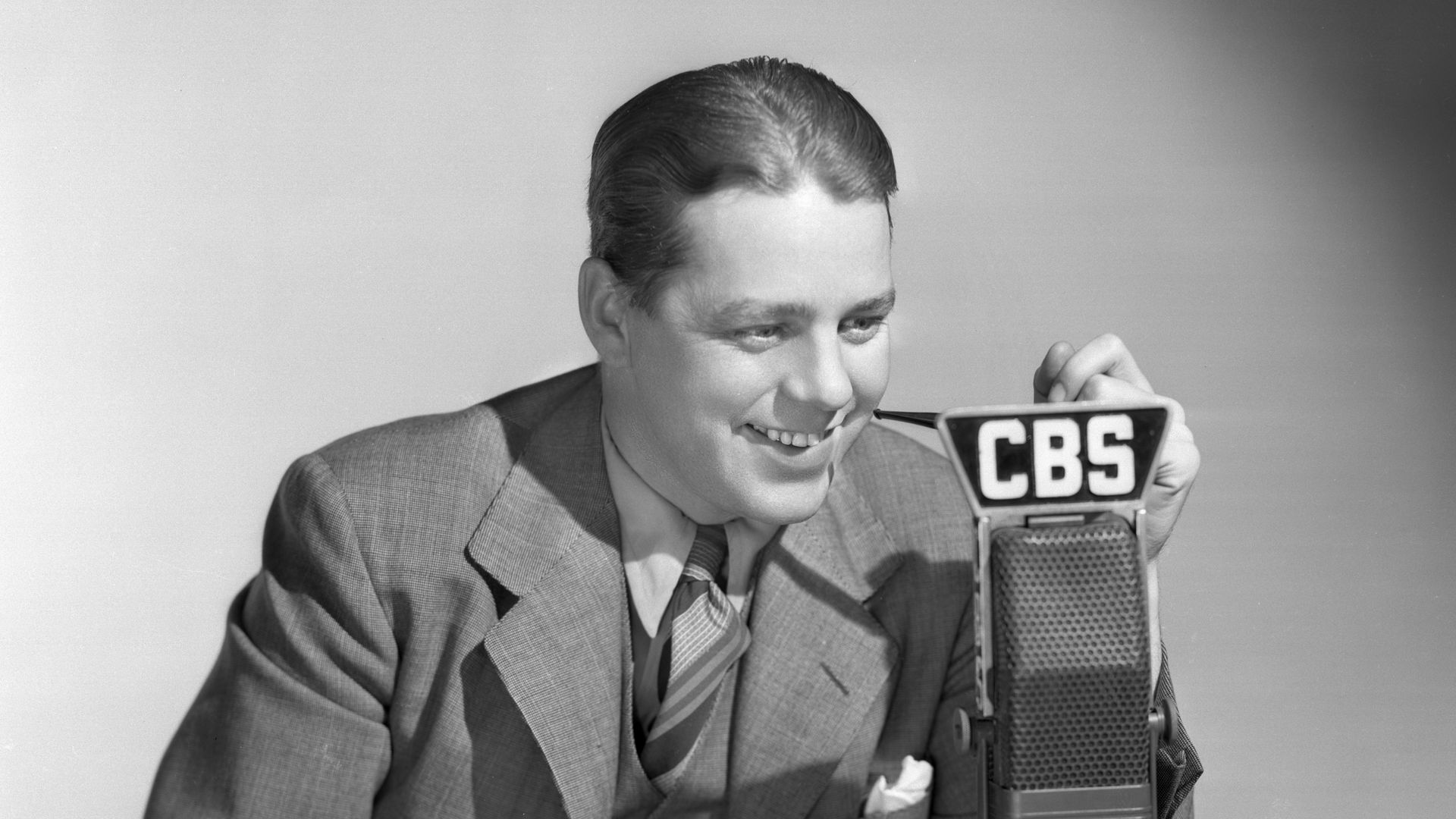 Black-and-white photo of a smiling man in a suit at a desk, speaking into a vintage CBS radio microphone. He holds papers; the desk has pencils, an eraser, and a notebook.