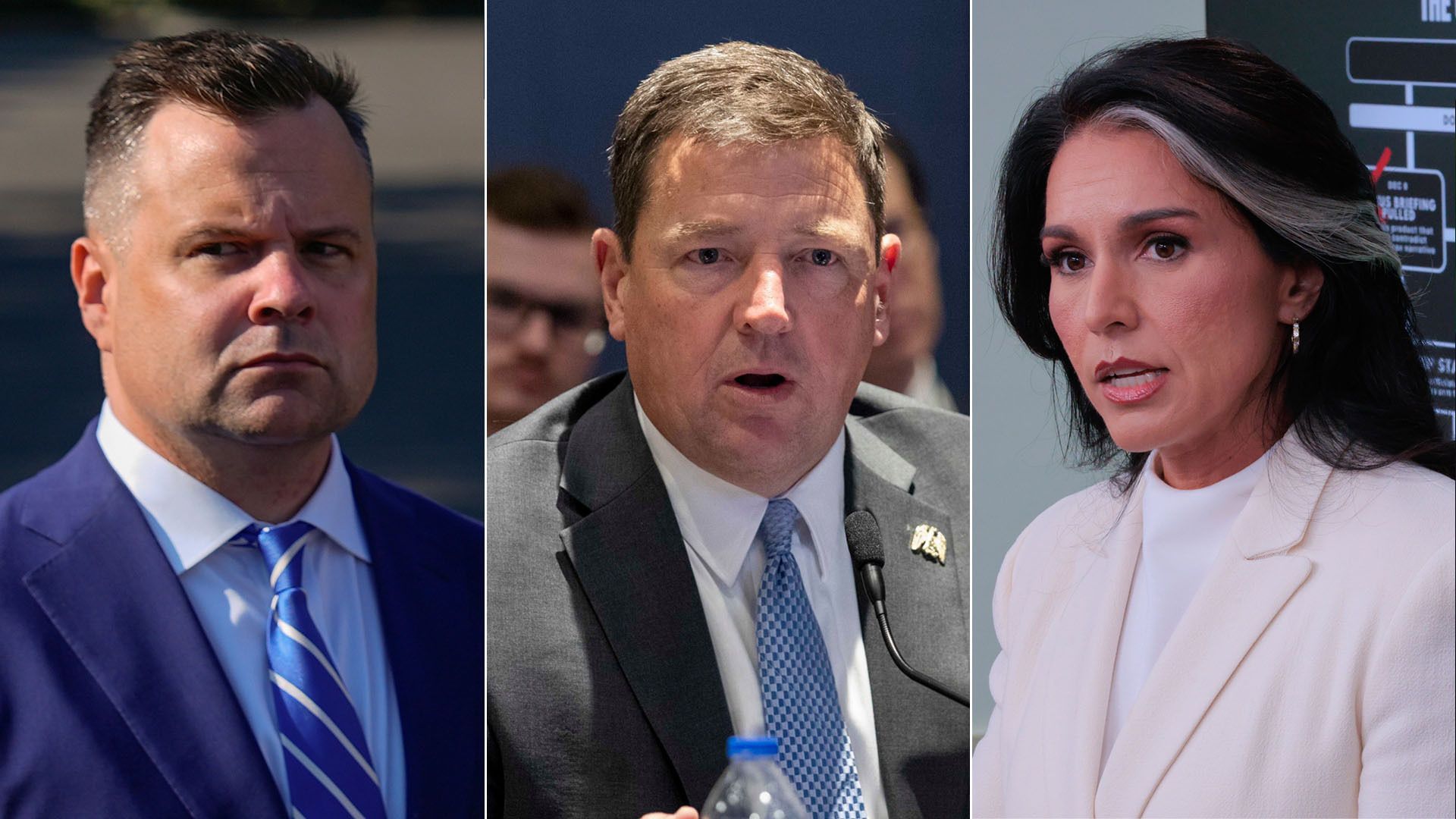 Three people in business attire: a man in a blue suit and striped tie looking serious, a man in a gray suit speaking into a microphone, and a woman in a white suit with dark hair and a white streak.