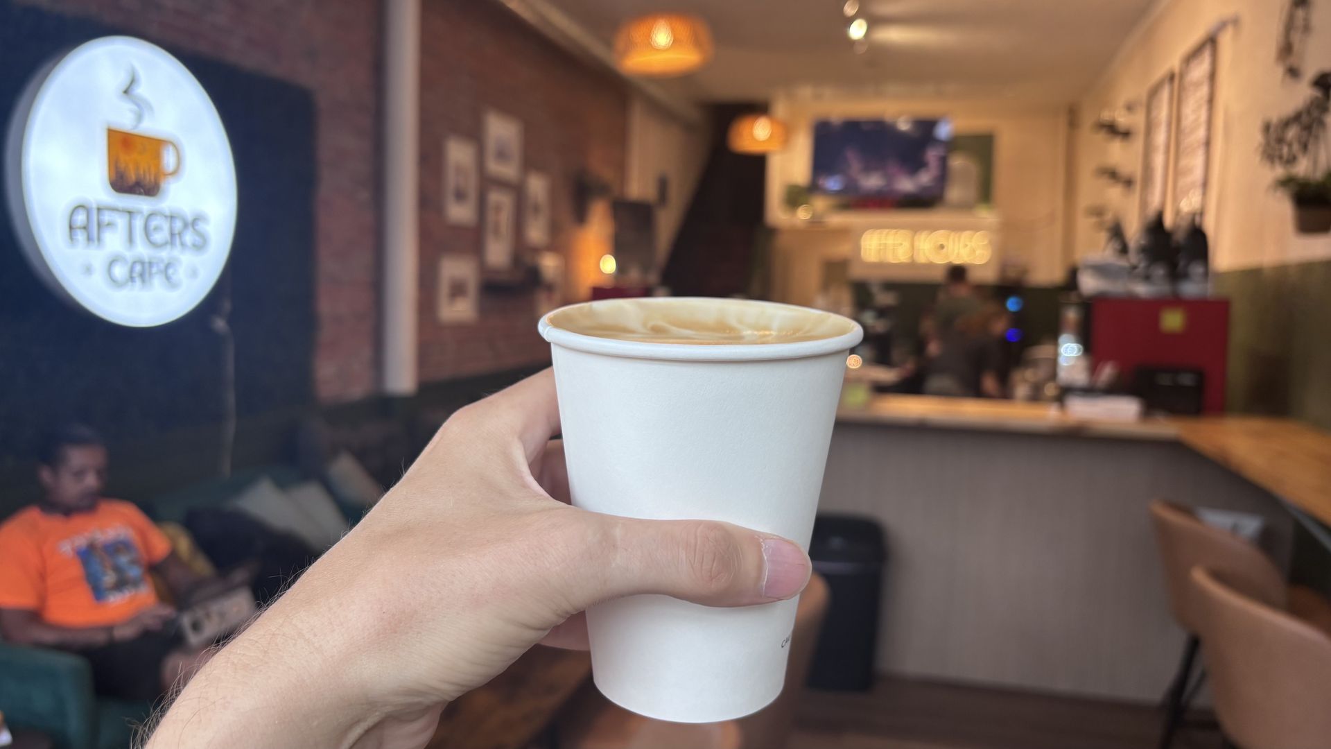 Hand holding a white paper coffee cup close to the camera inside a cozy cafe with brick walls, framed art, and warm pendant lights, while blurred patrons sit in the background.