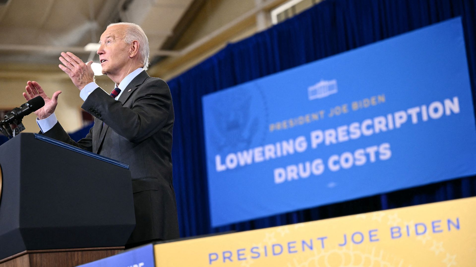 Joe Biden speaks at a lectern. A poster behind him reads "lowering prescription drug costs." 