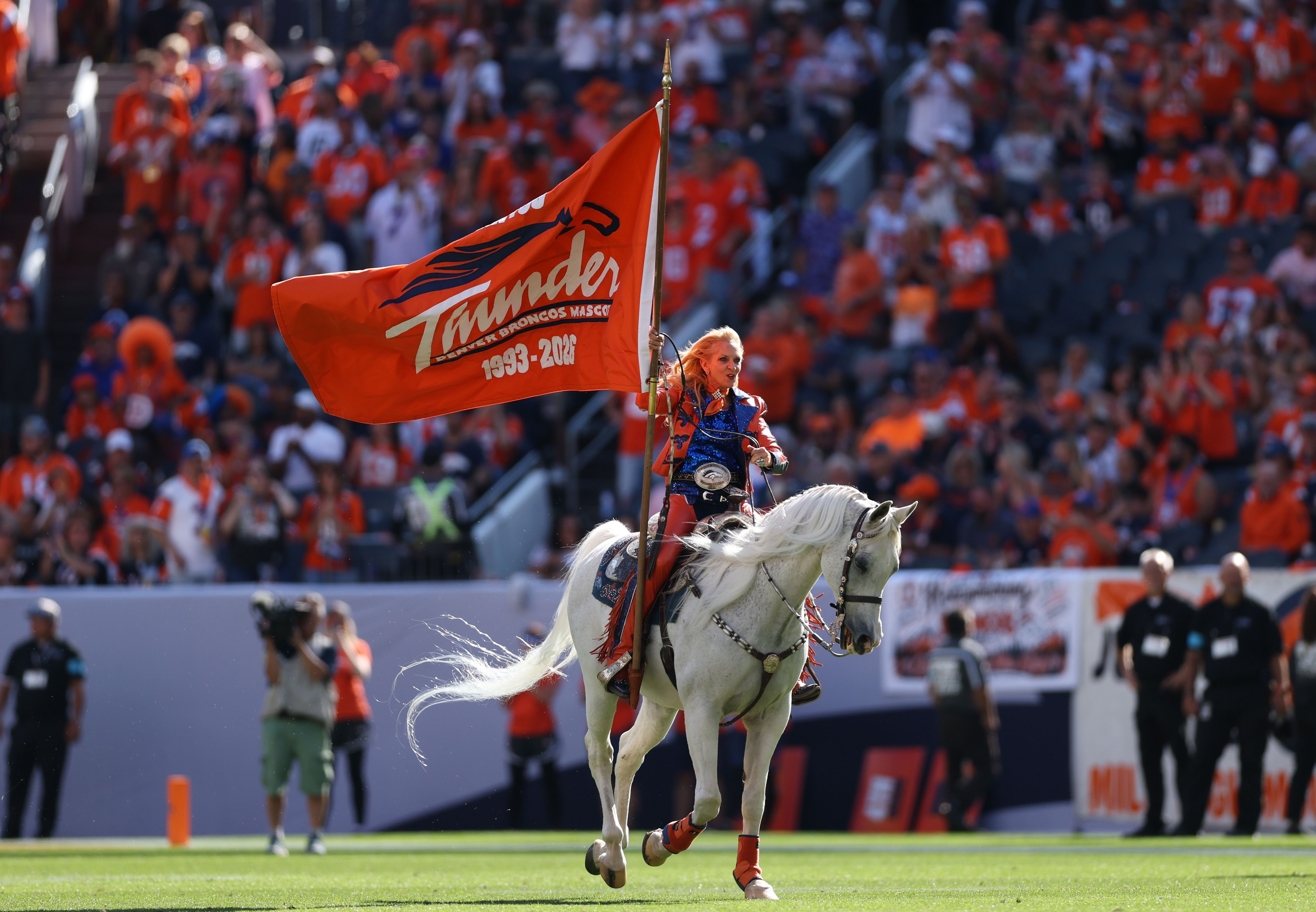 An equestrian rider on a white horse holds a tall orange flag with the white script "Thunder" and "Denver Broncos Mascot" text, as a large crowd in orange fills a stadium.