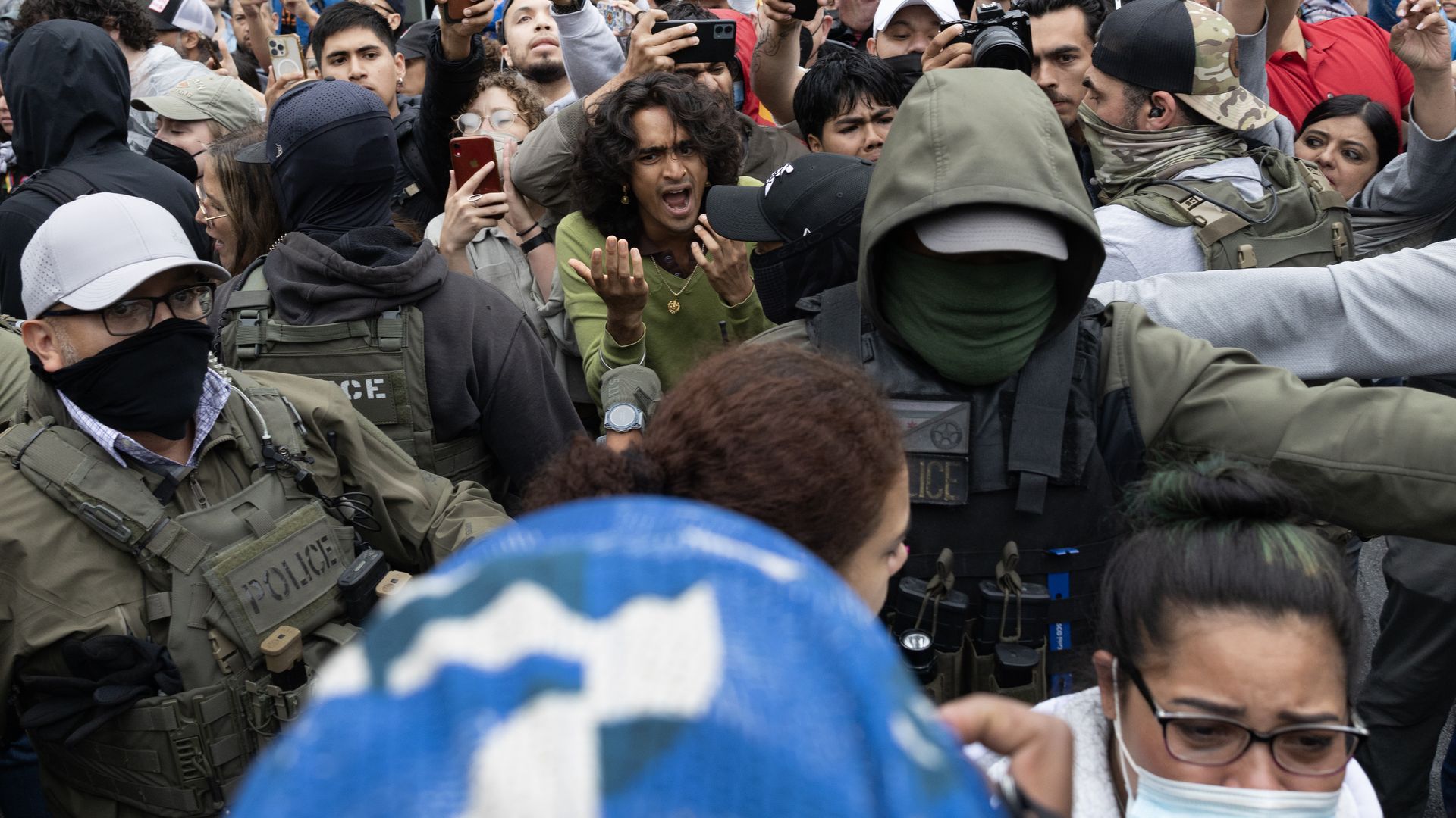 Photo of a crowd pushing against officers as they escort people into a van. 
