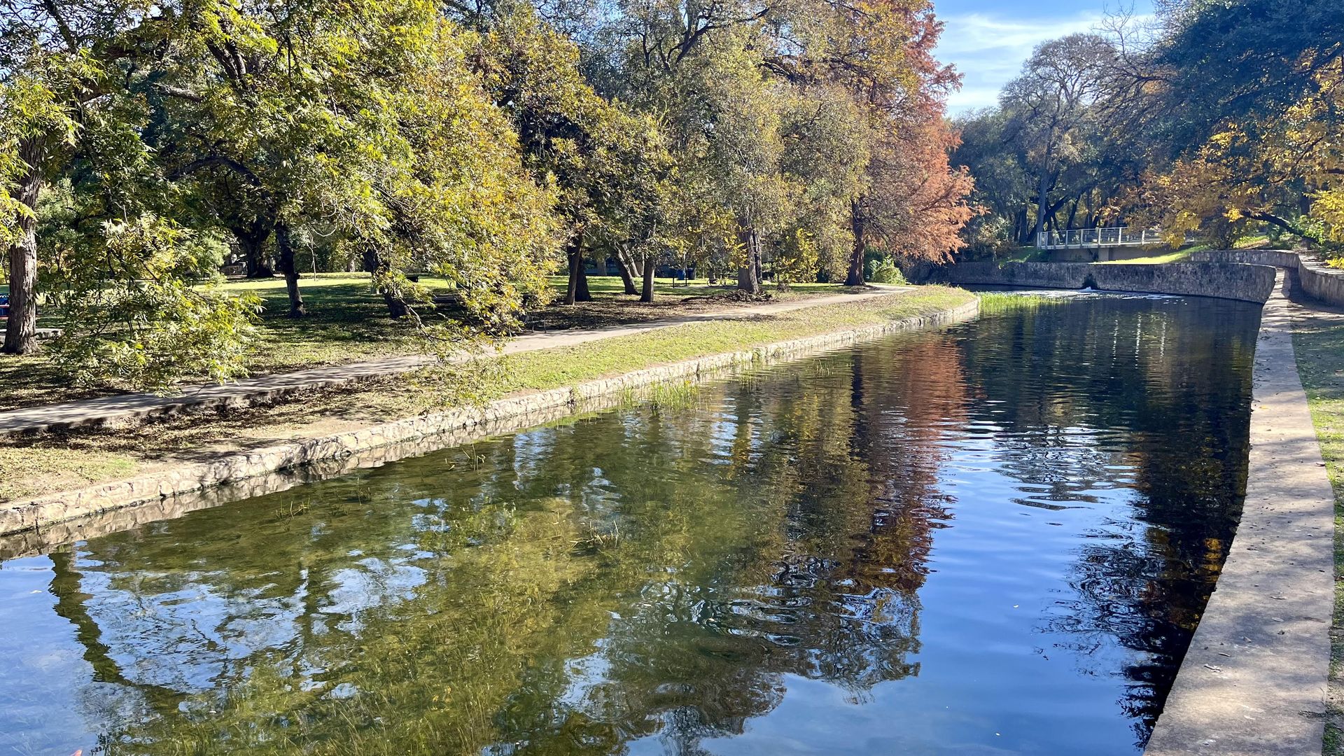 The San Antonio River, with its retaining walls, runs through trees in Brackenridge Park that are reflected in the water.