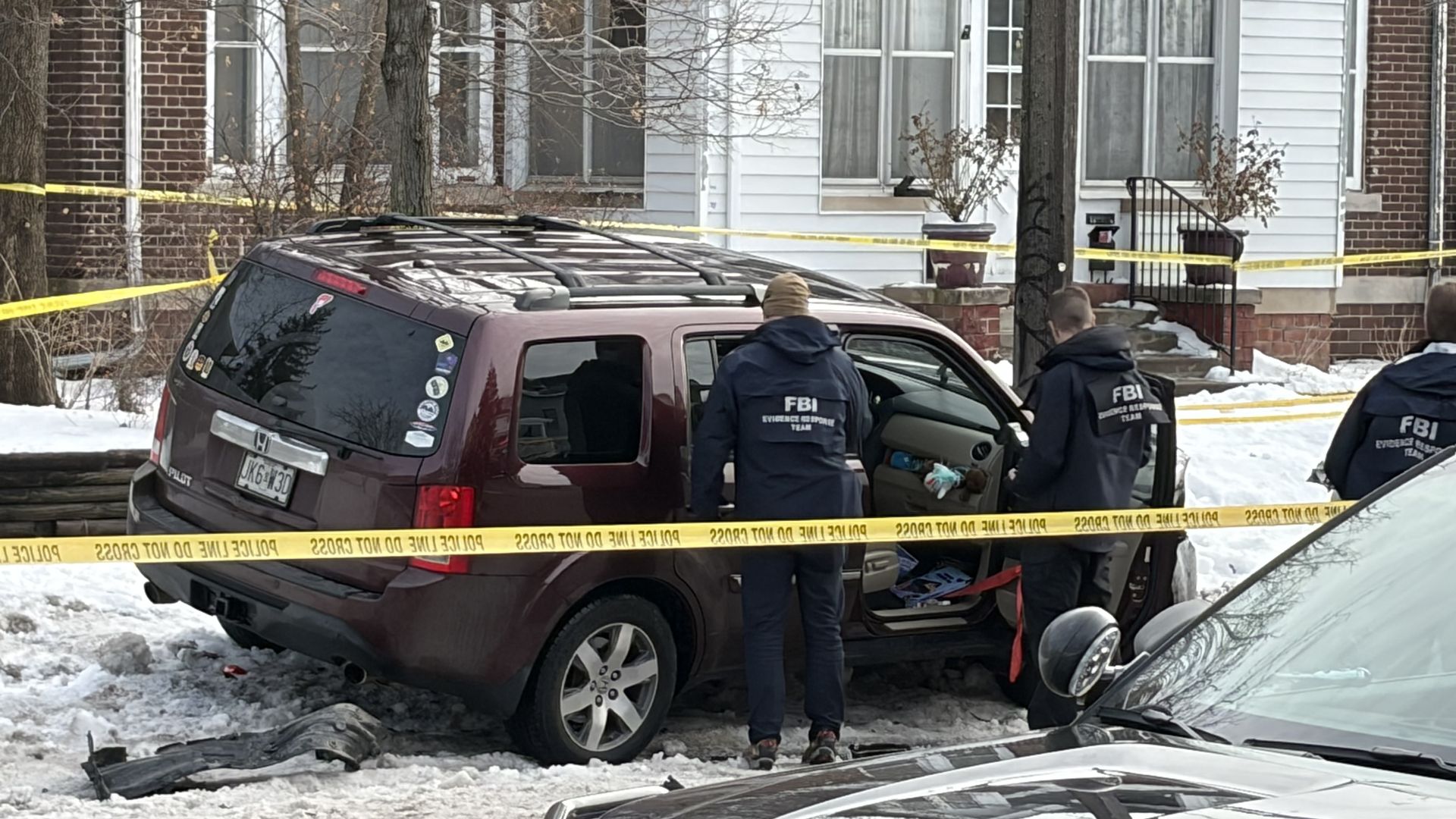 Three FBI Evidence Response Team agents inspecting a maroon Honda Pilot SUV with broken windows, surrounded by yellow police tape on a snowy street in front of houses.