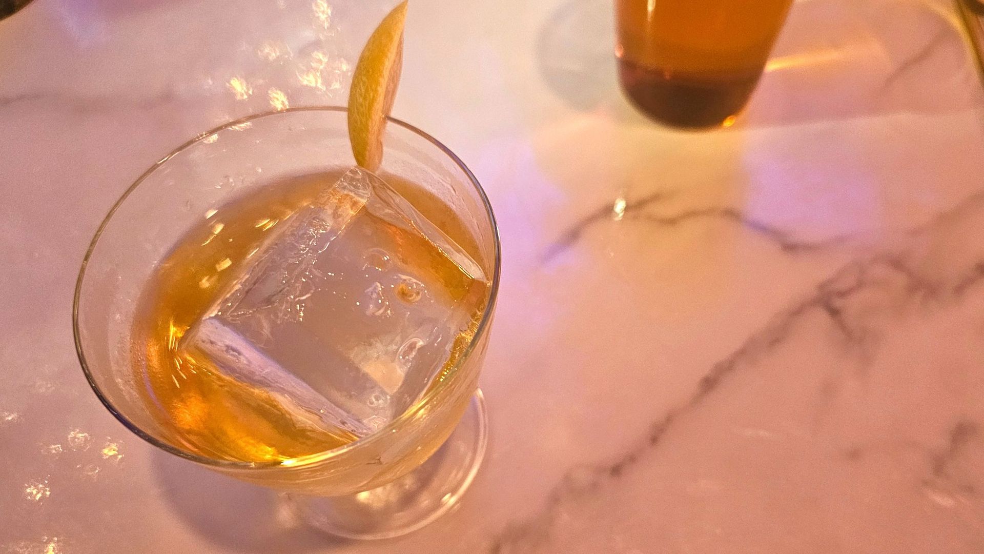 Close-up of a glass with amber liquid, a large clear ice cube, and a lemon peel garnish on a marble surface with warm lighting.