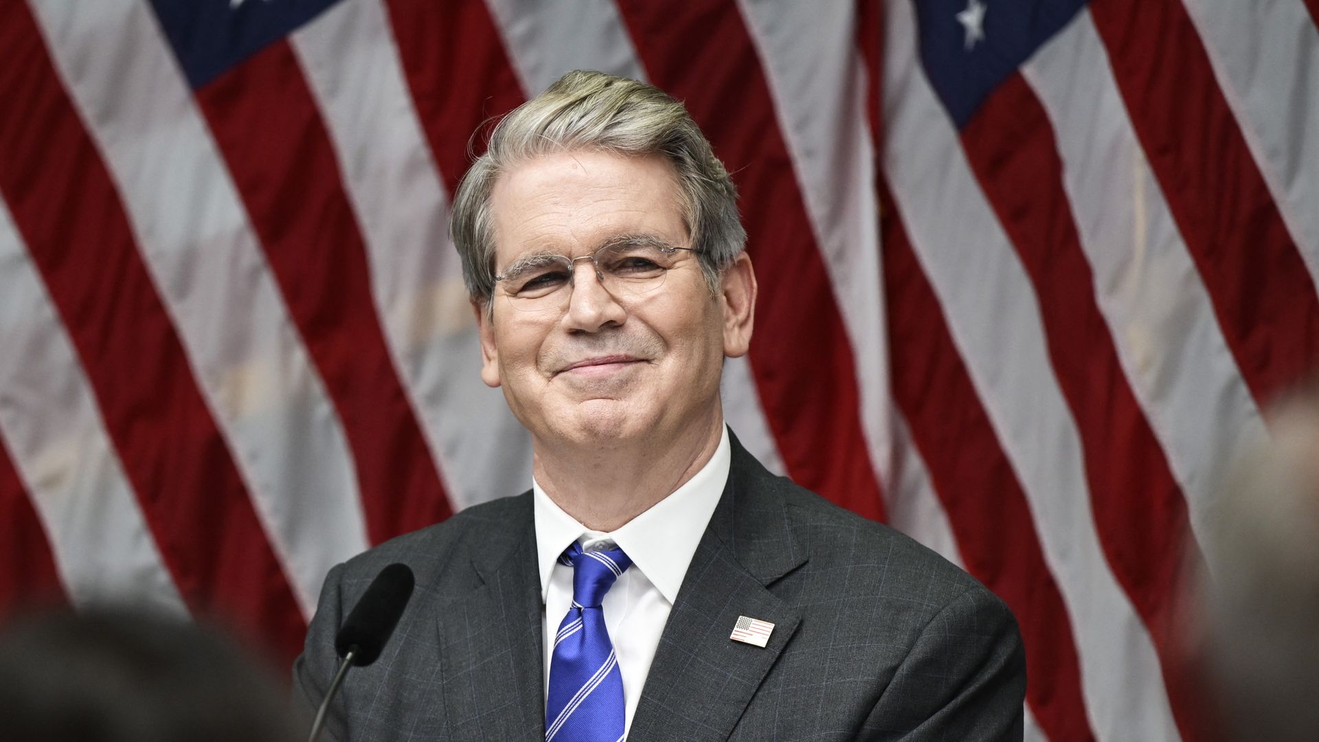 Treasury Secretary Scott Bessent in a dark suit and blue striped tie with an American flag pin, standing at a microphone with multiple U.S. flags in the background.