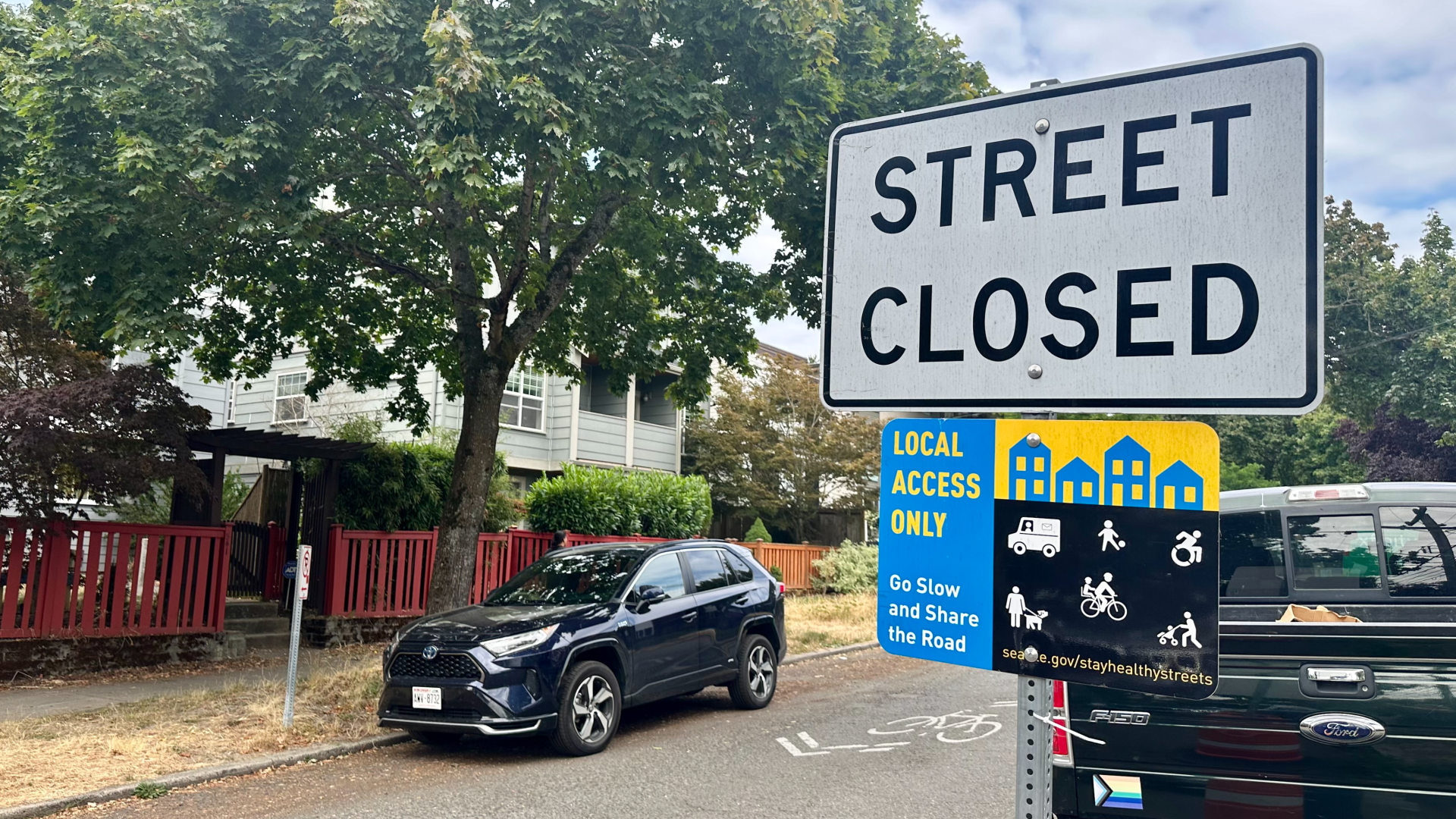 A view of a street with a large tree in background and two black cars parked along the curb, with a sign that says "Street Closed" and "Local Access Only" in the foreground.