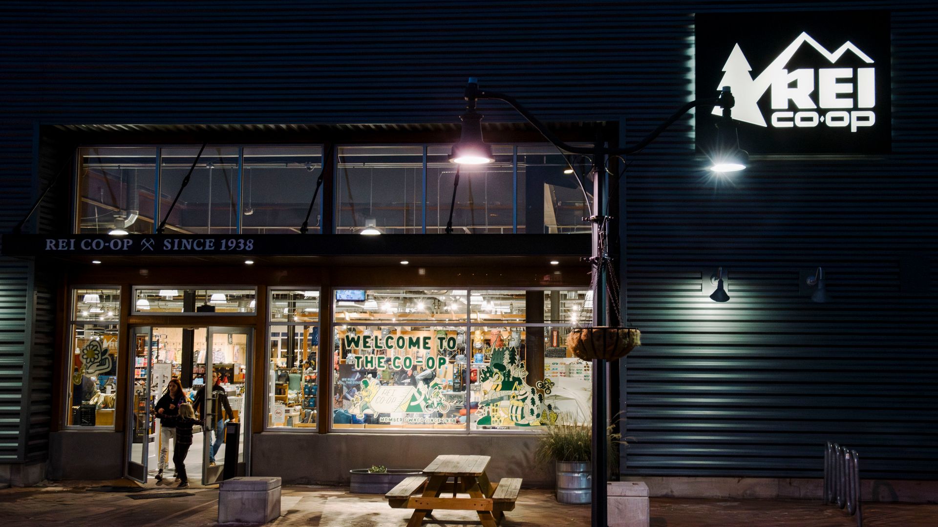 Two people walk at night into the front door of an REI store, with the interior lit brightly and paint in the window that says "welcome to the co-op."
