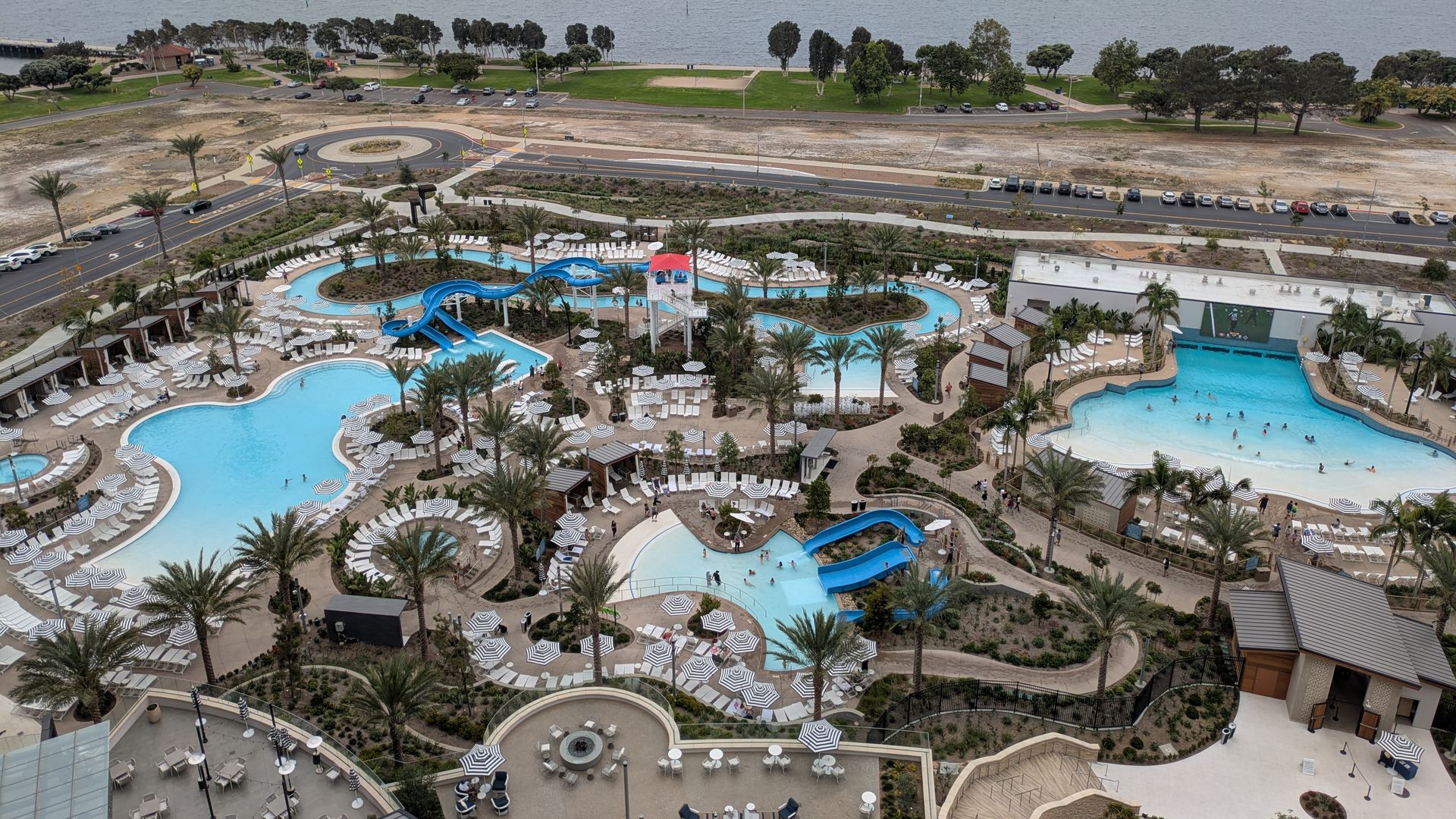 Aerial view of a resort pool with blue slides and many white chairs, palm trees and green lawns, a roundabout and road, gazing over a calm bay with distant buildings on the horizon.