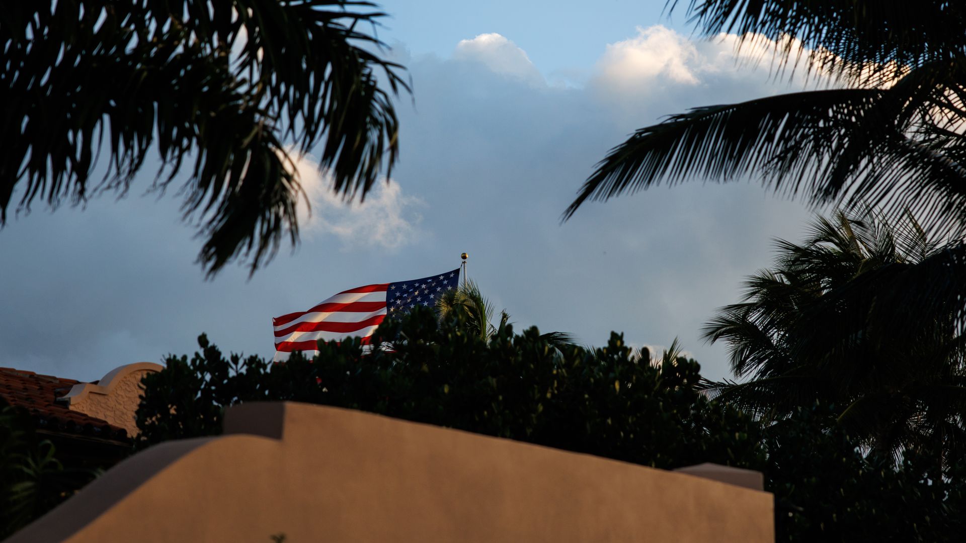 An American flag can be seen in the sky poking above a wall and a treeline. Palm trees can be seen in the foreground.