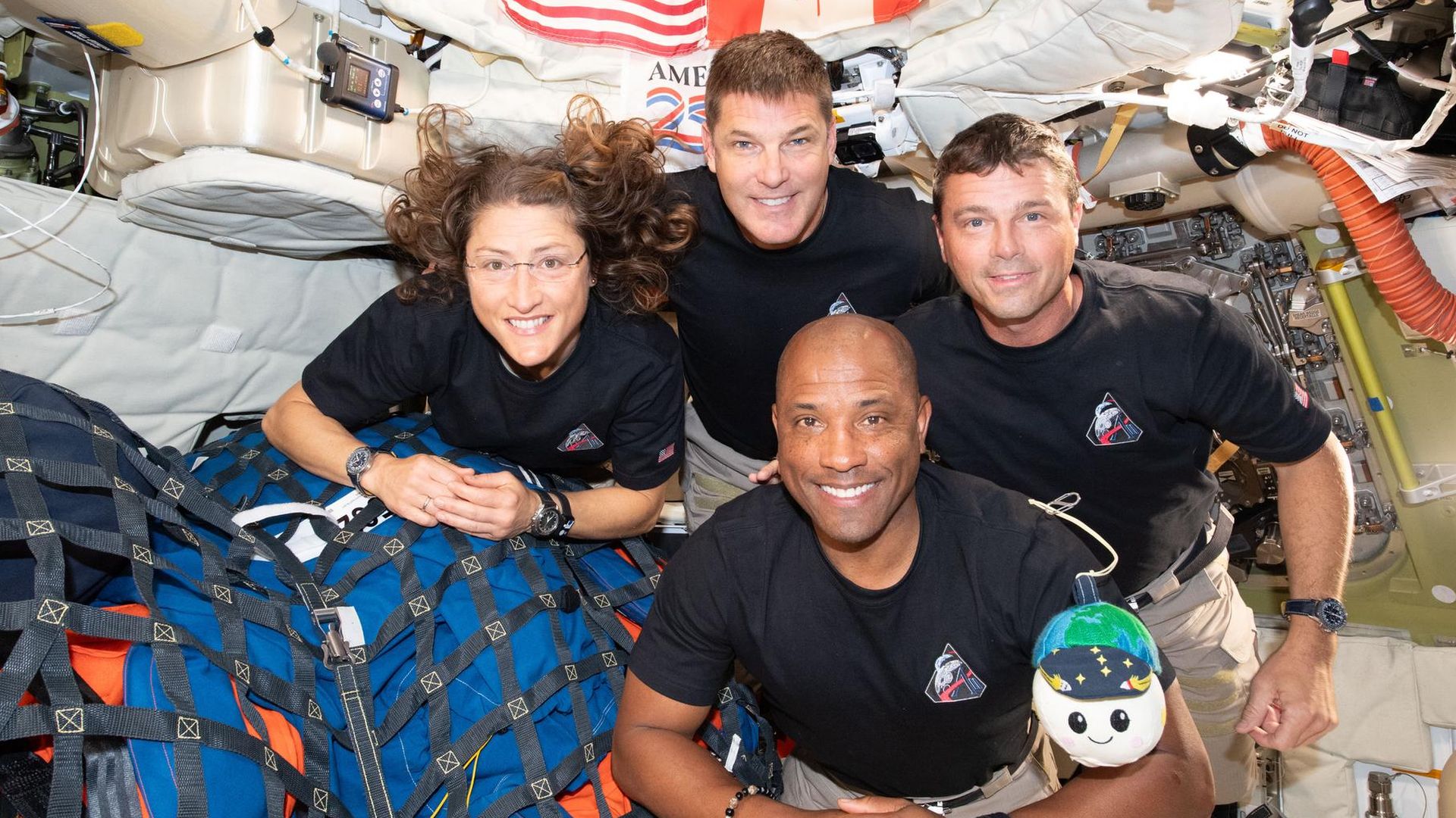 Four astronauts in black shirts float inside a space station module, smiling for a group photo. US and Canadian flags hang overhead amid cables and gear; one crew member holds a small plush toy.